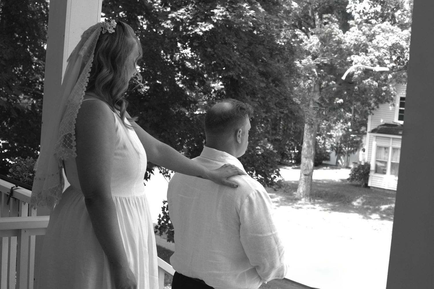 Black and white photo of a bride in veil and groom kneeling on a porch, sharing a romantic wedding moment.