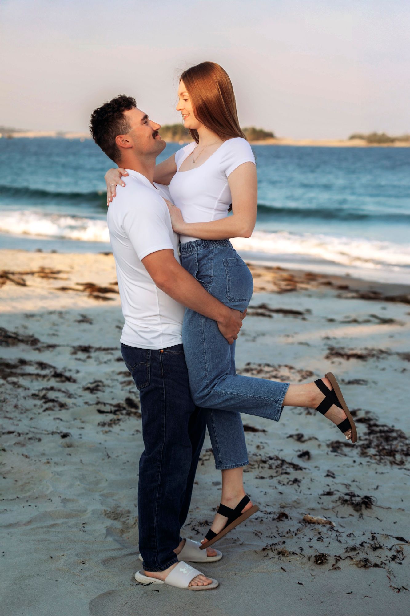 Couple in matching white shirts and jeans embracing on a sandy beach with ocean waves in the background.