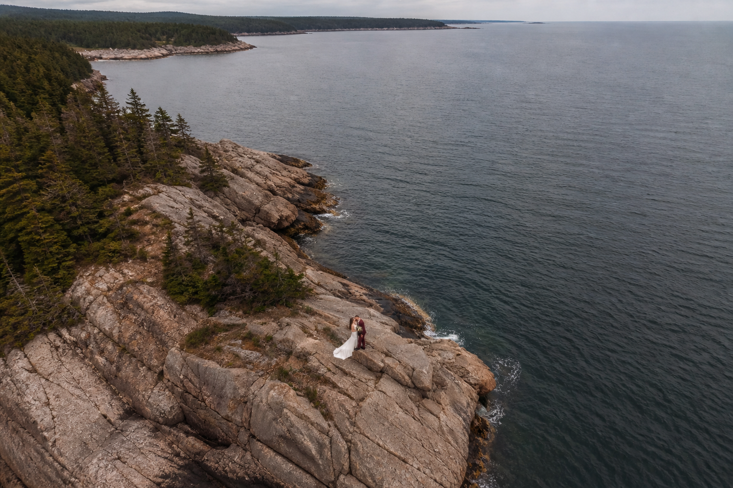 Halifax ns Aerial view of a couple in wedding attire standing on rocky coastal cliffs overlooking the ocean, surrounded by evergreen trees.