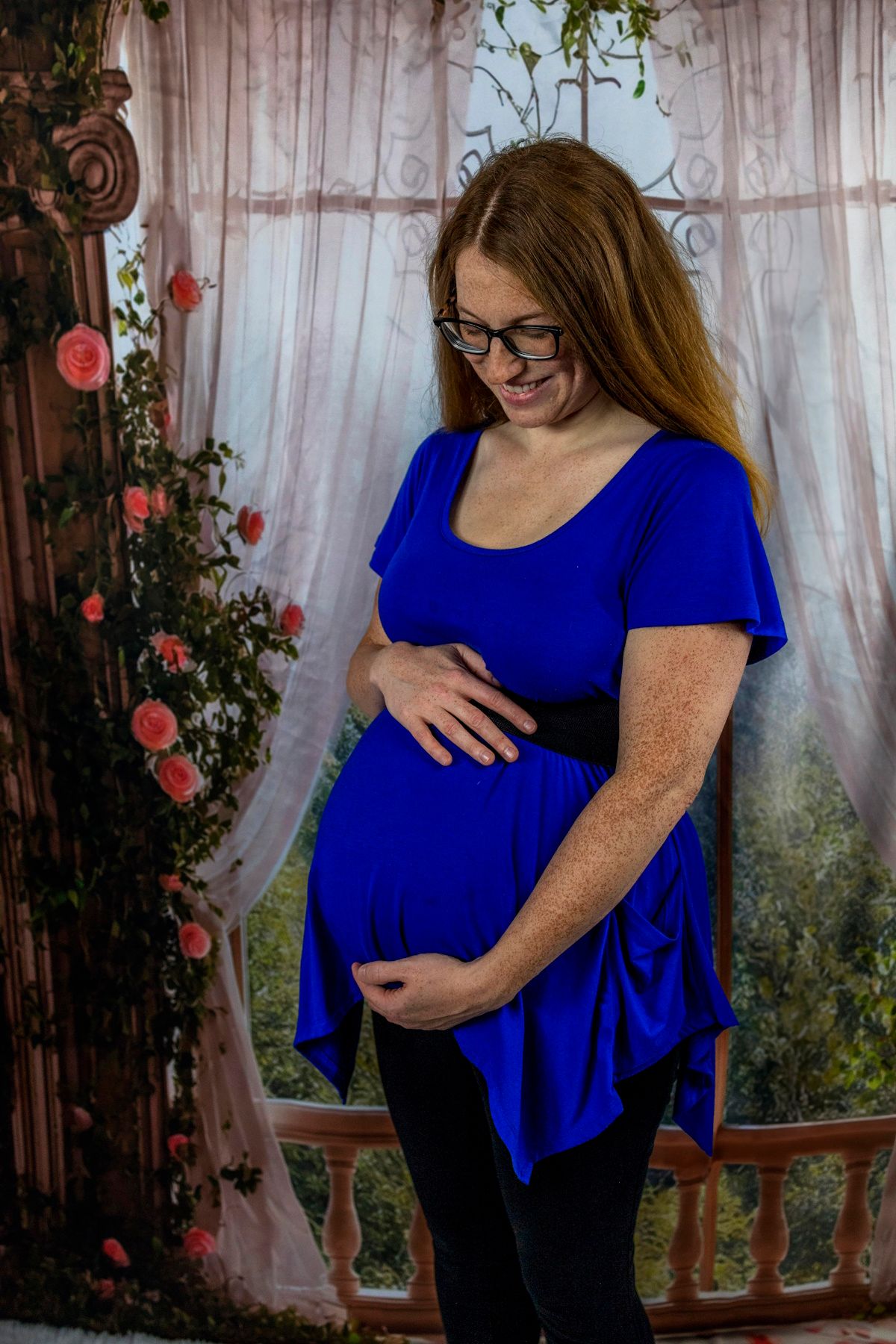 Pregnant woman in blue top cradling baby bump, wearing glasses, standing before floral backdrop with pink roses.