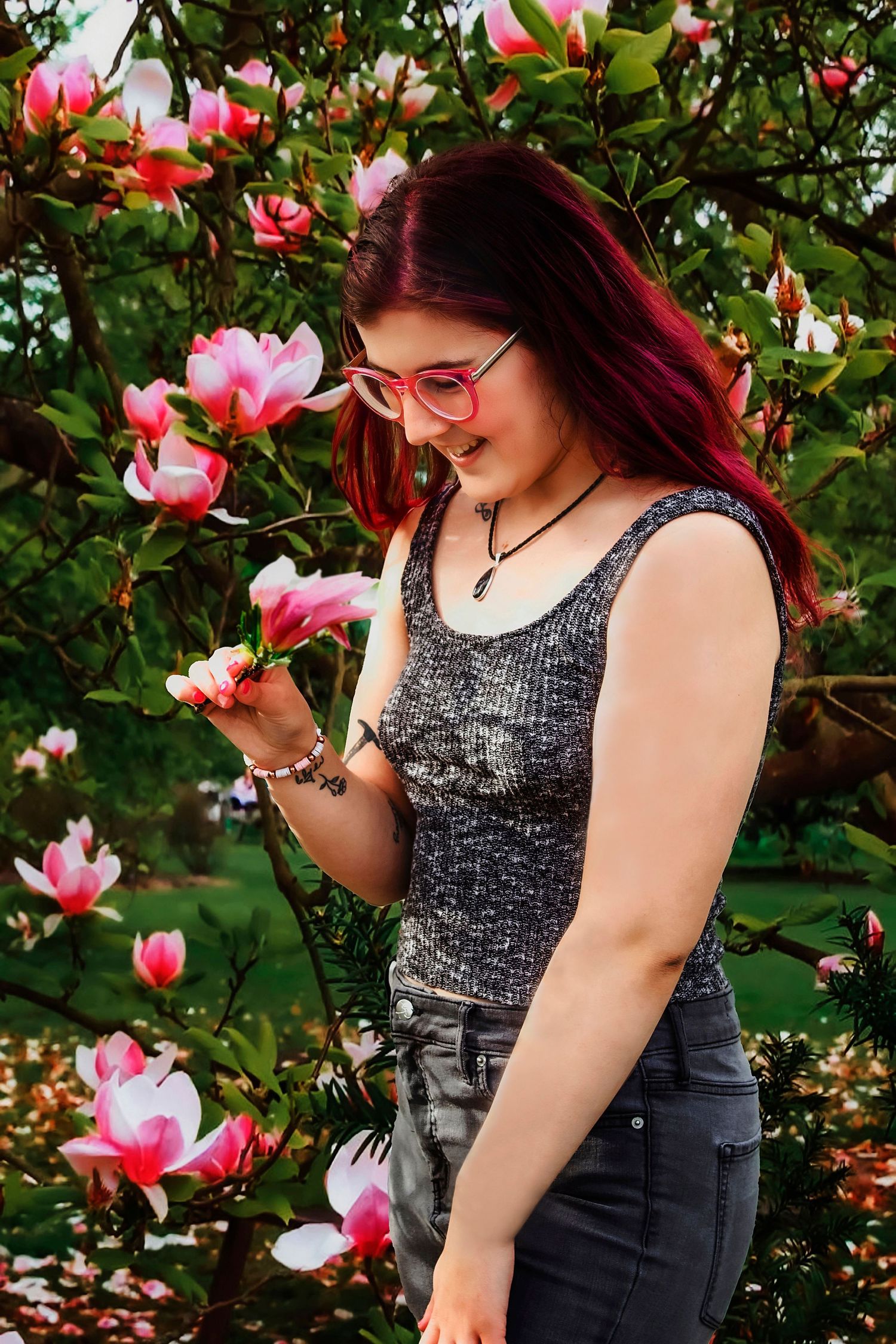 Young woman with red hair and pink glasses smelling pink magnolia blossoms in a lush green garden.
