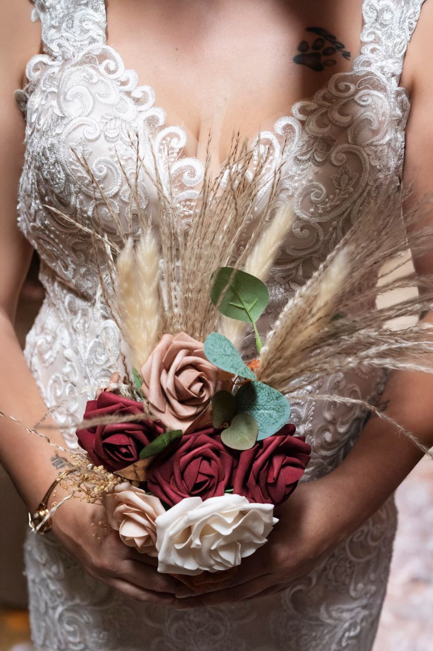 Bride in lace wedding dress holding a boho bouquet with red, pink, white roses and dried pampas grass.