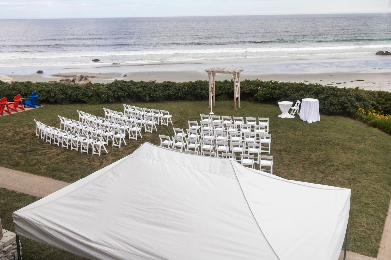 Outdoor beachfront wedding ceremony setup with white chairs, wooden arch, and ocean views on a sunny day.