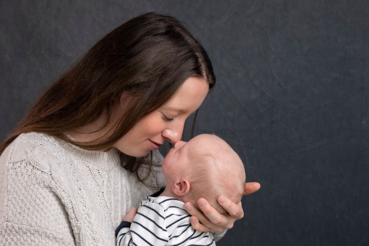 in Chester N.S A mother tenderly holds her newborn baby close, touching foreheads in an intimate moment against a dark gray background.