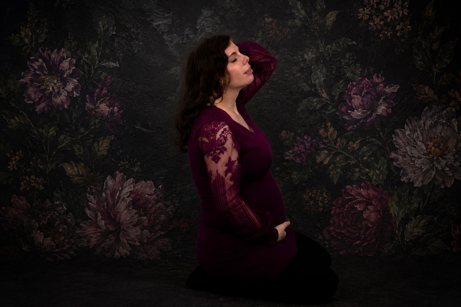 at our South Shore, studio a Pregnant woman in burgundy lace gown kneeling gracefully against dark floral backdrop in maternity portrait.