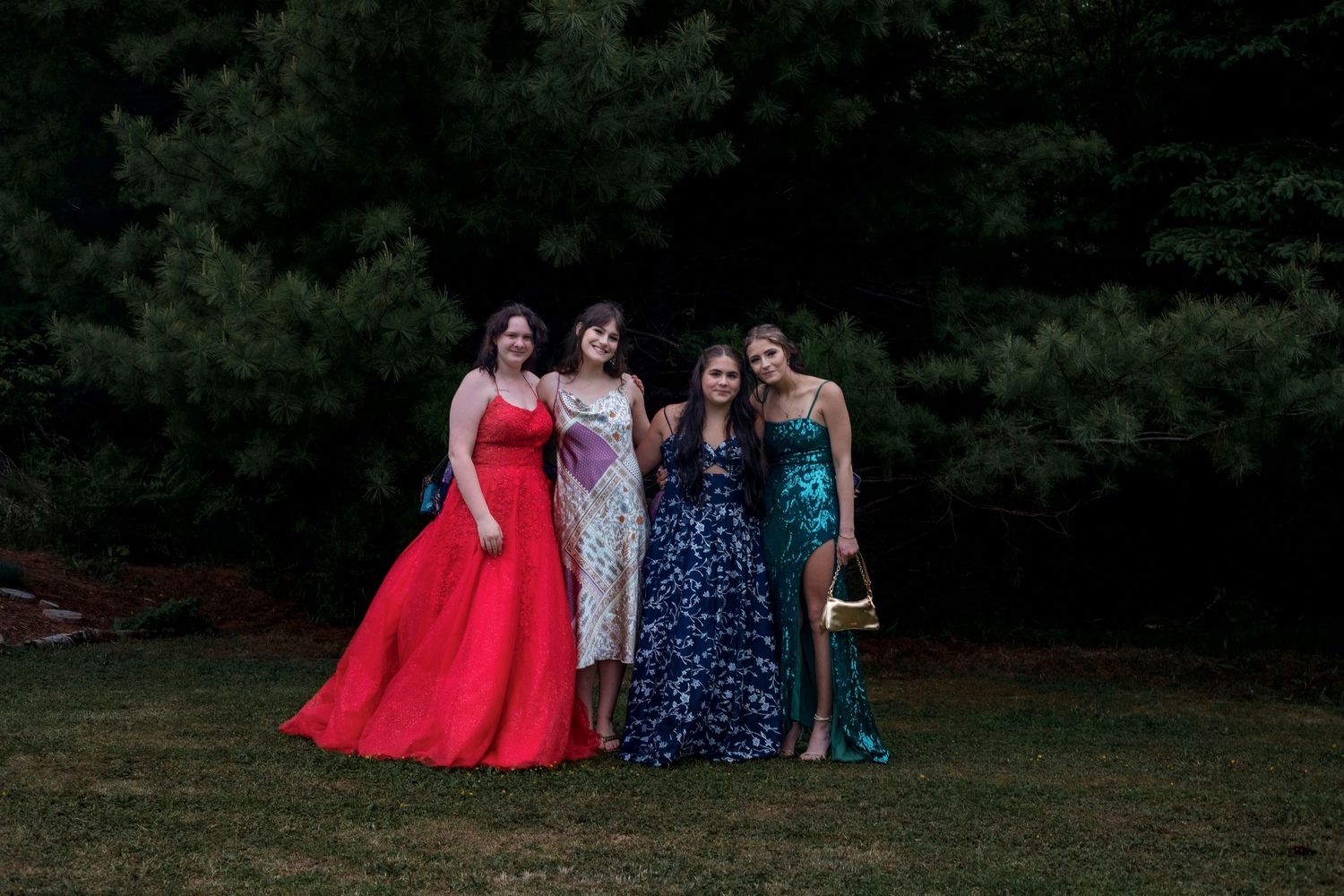 Four young women in formal prom gowns posing together outdoors in front of evergreen trees.