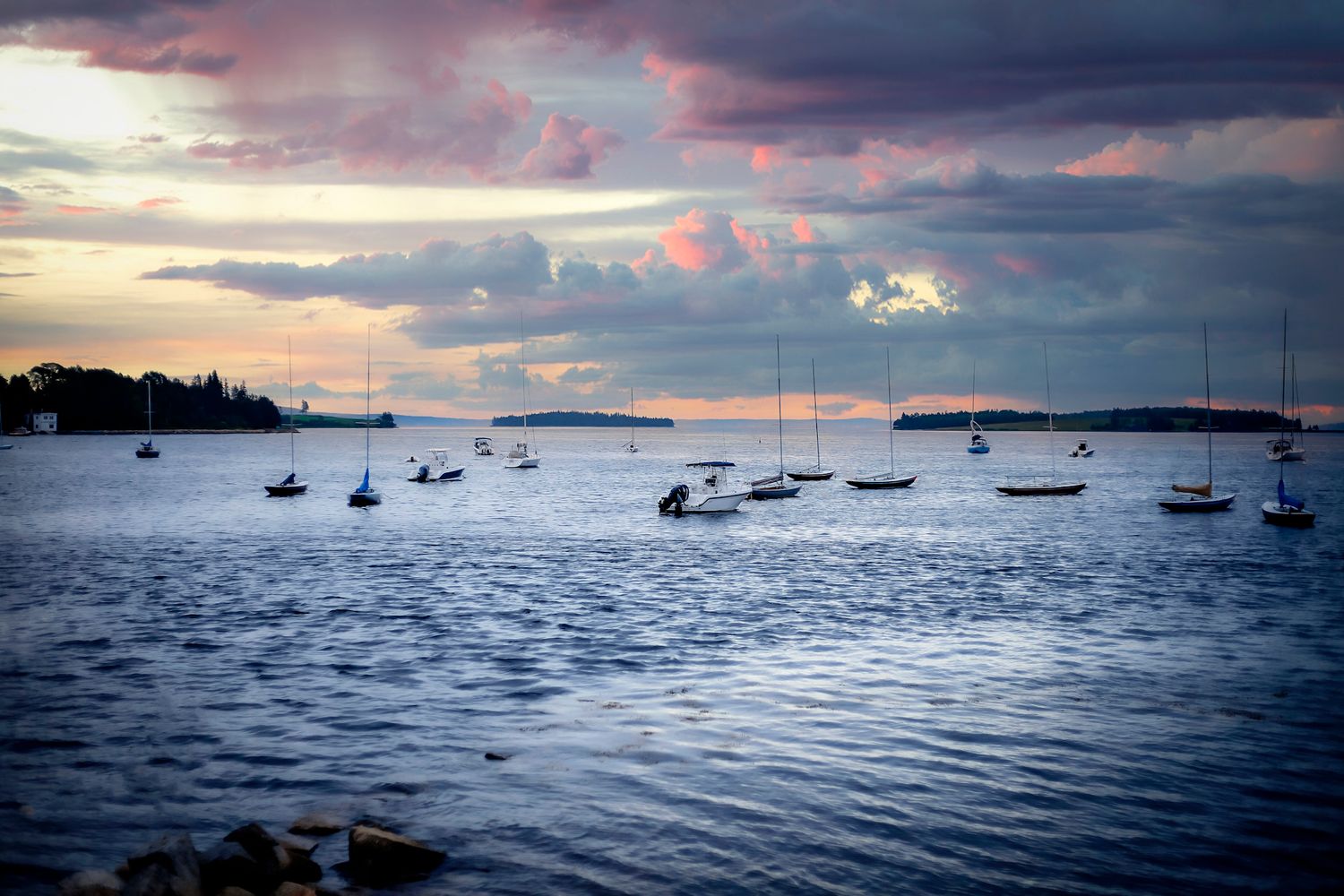 Sailboats moored in a calm harbor at sunset with dramatic cloudy sky and silhouetted islands in the background.