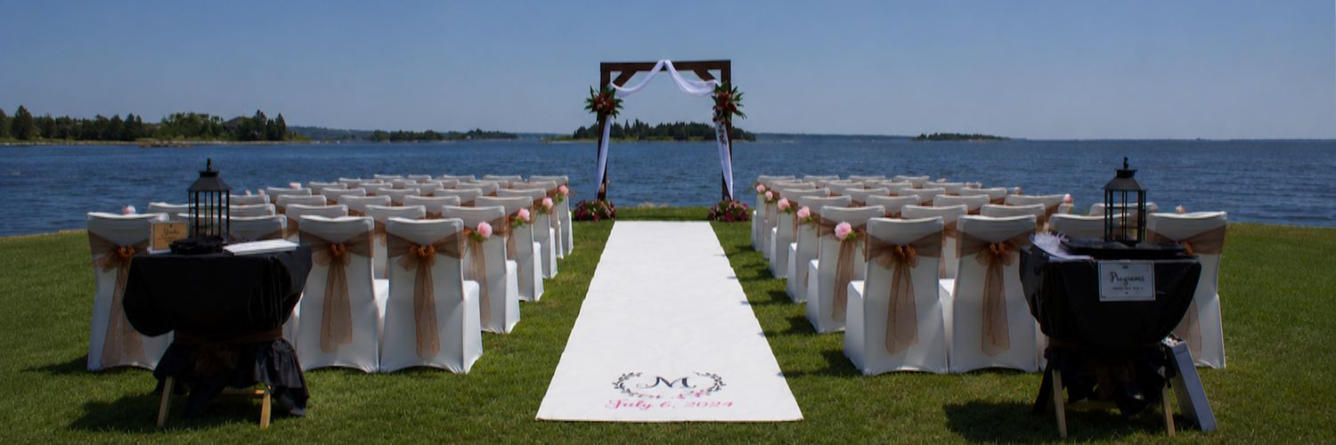 Elegant outdoor waterfront wedding ceremony setup with white aisle runner, draped chairs, and a rustic wooden arch.