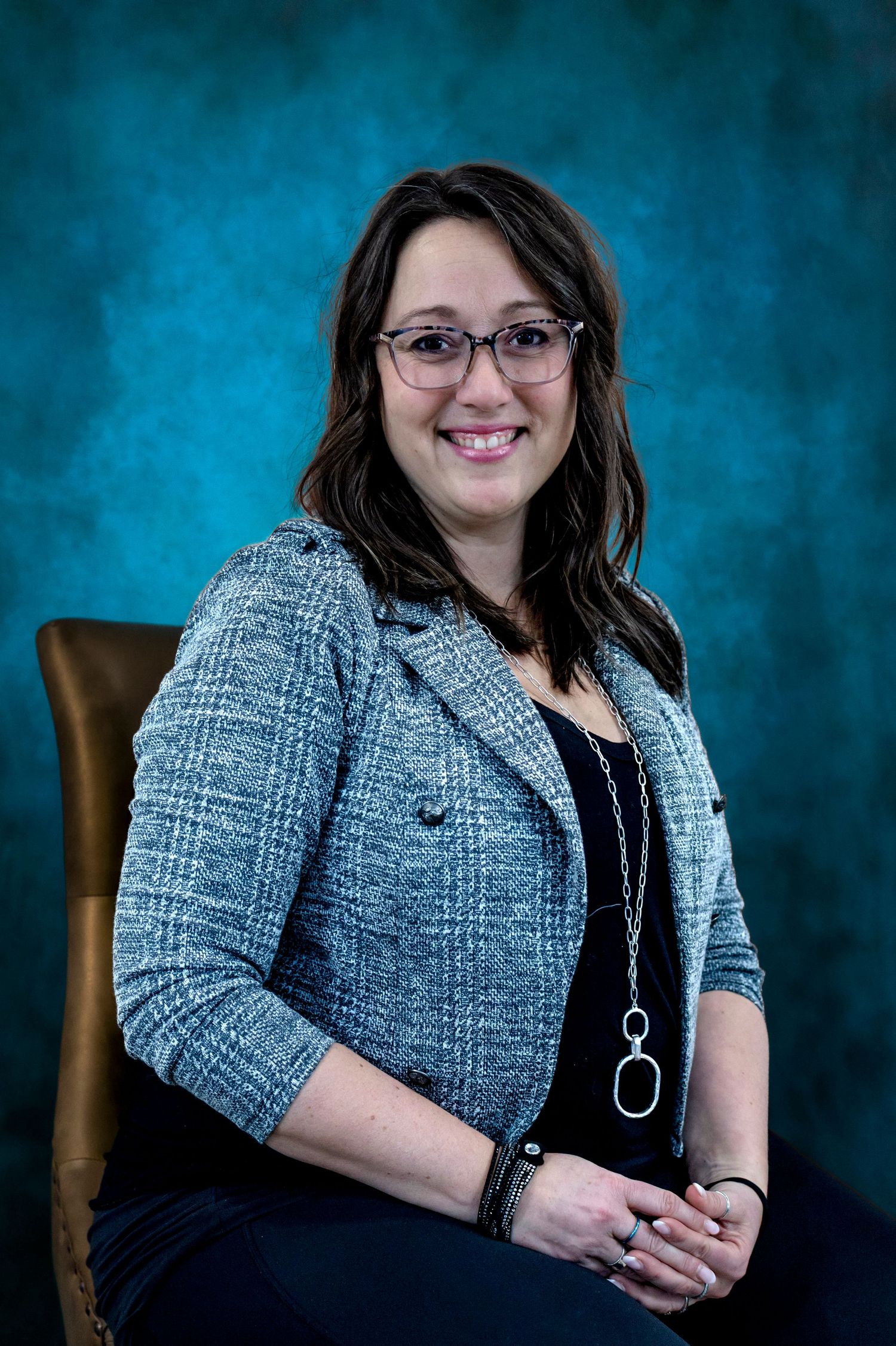 A smiling woman with glasses in a gray tweed blazer sits in a chair against a teal studio background.