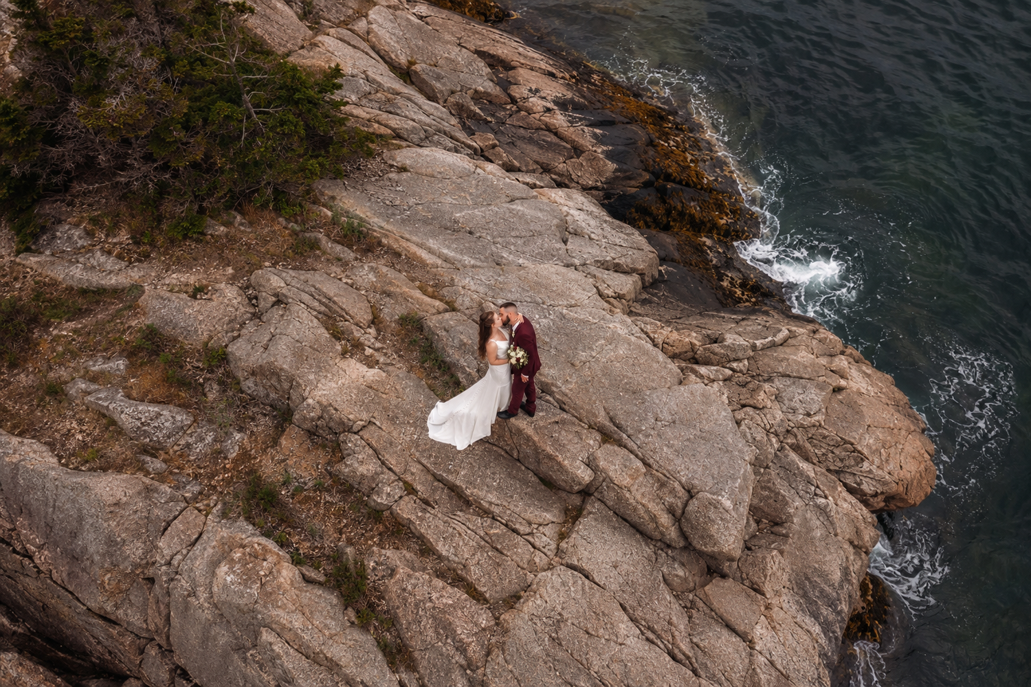 Aerial view of a bride and groom posing on rocky coastal cliffs with ocean waves below on their wedding day.