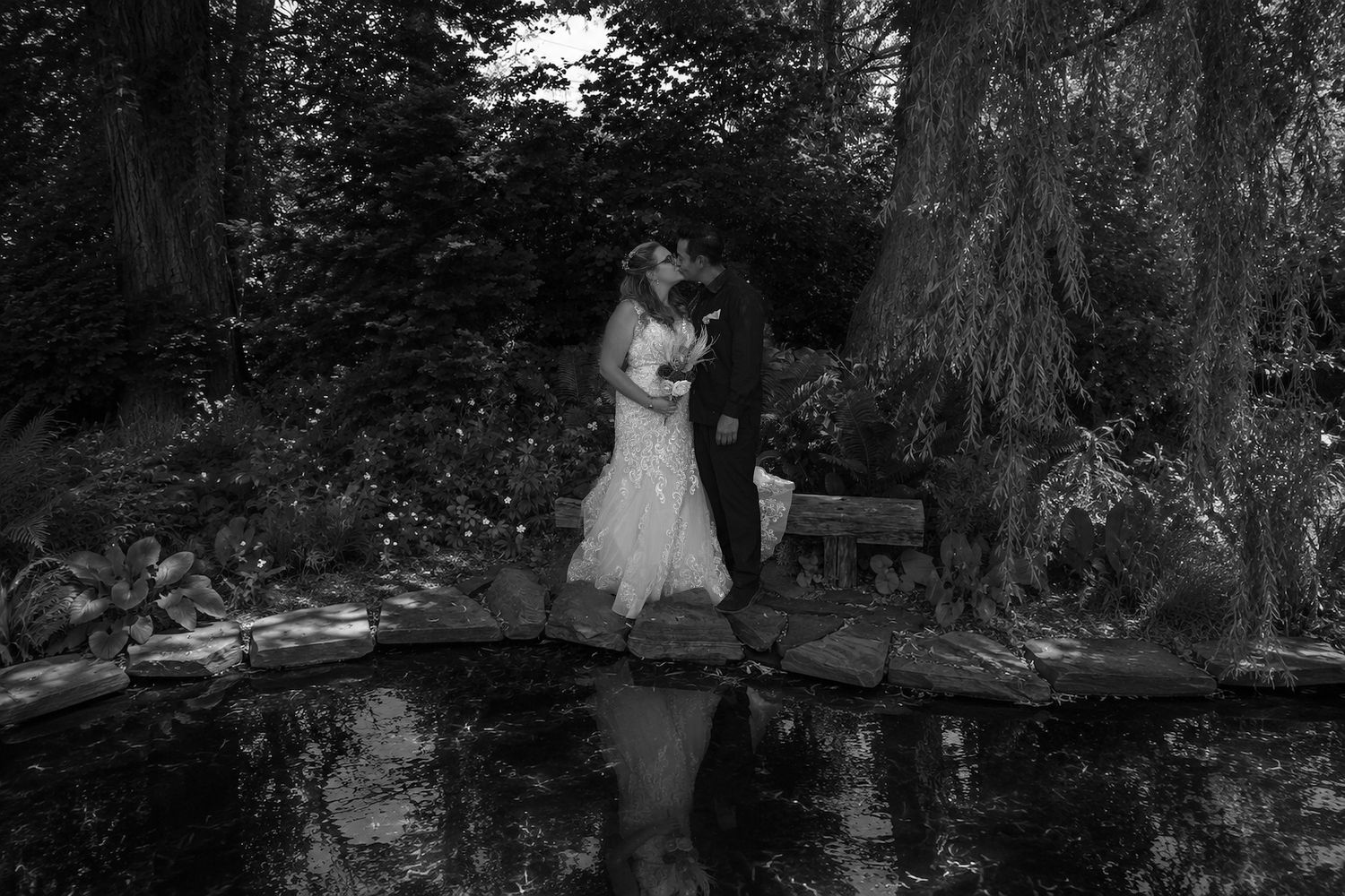 Black and white photo of a bride and groom kissing on a bridge over a pond surrounded by lush garden greenery.
