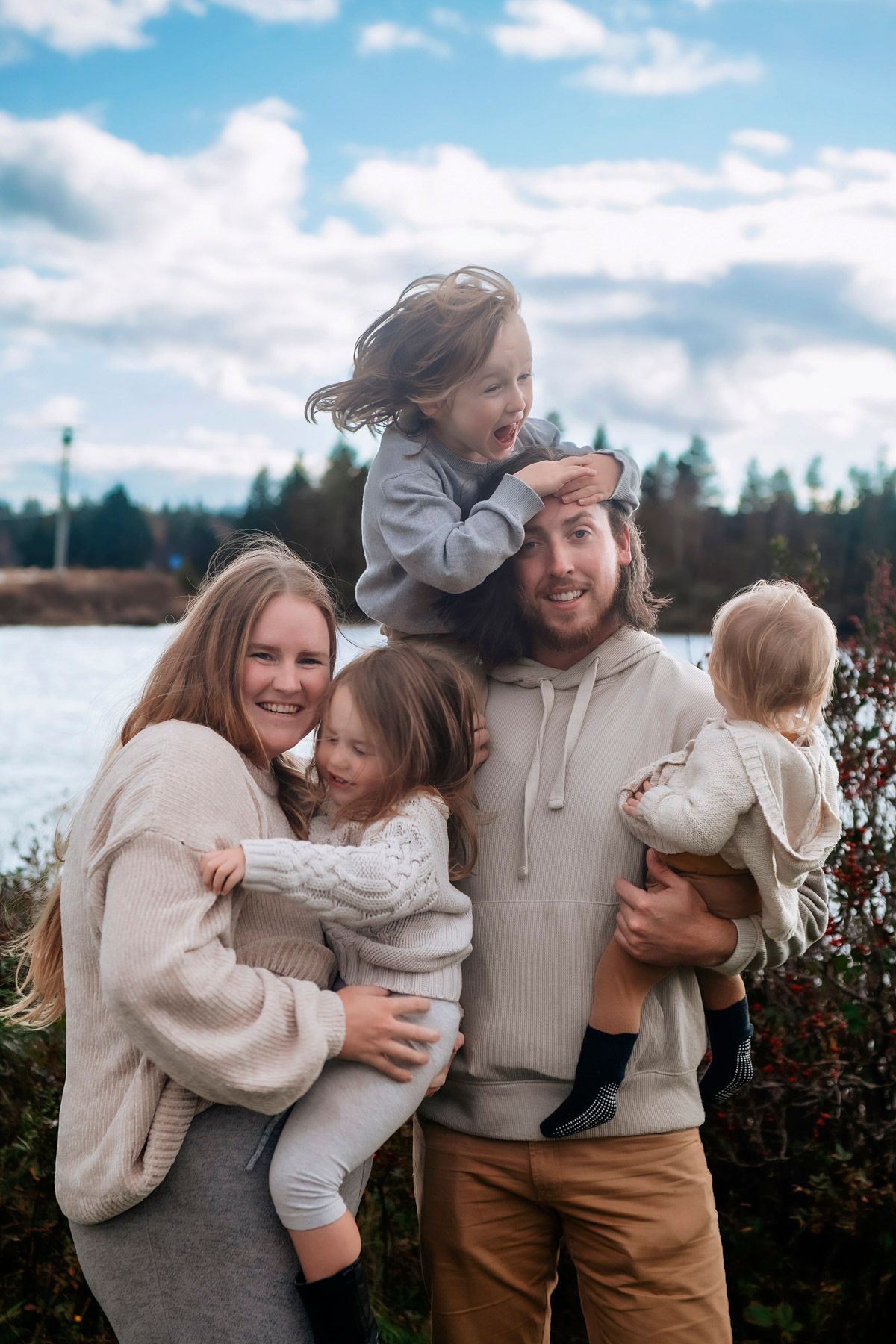Happy family of five posing outdoors near a lake with evergreen trees and cloudy sky in the background.