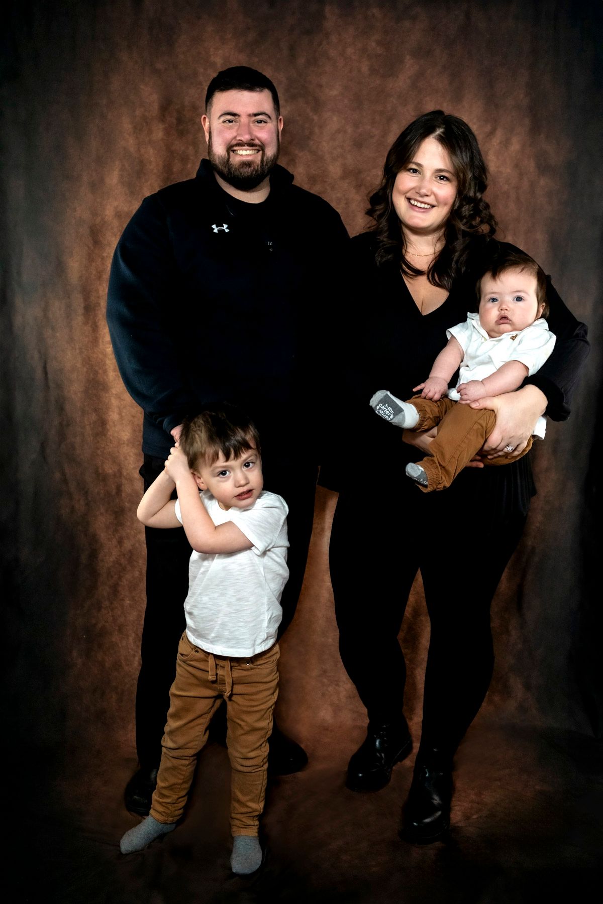 Annapolis Valley Family portrait of parents with toddler son and baby infant posing together against brown studio backdrop.
