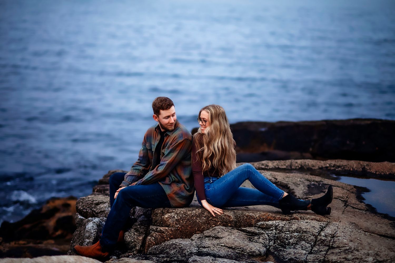 in Halifax a Couple sitting on rocky coastal shore with ocean backdrop, wearing casual fall outfits, sharing an intimate engagement shoot.