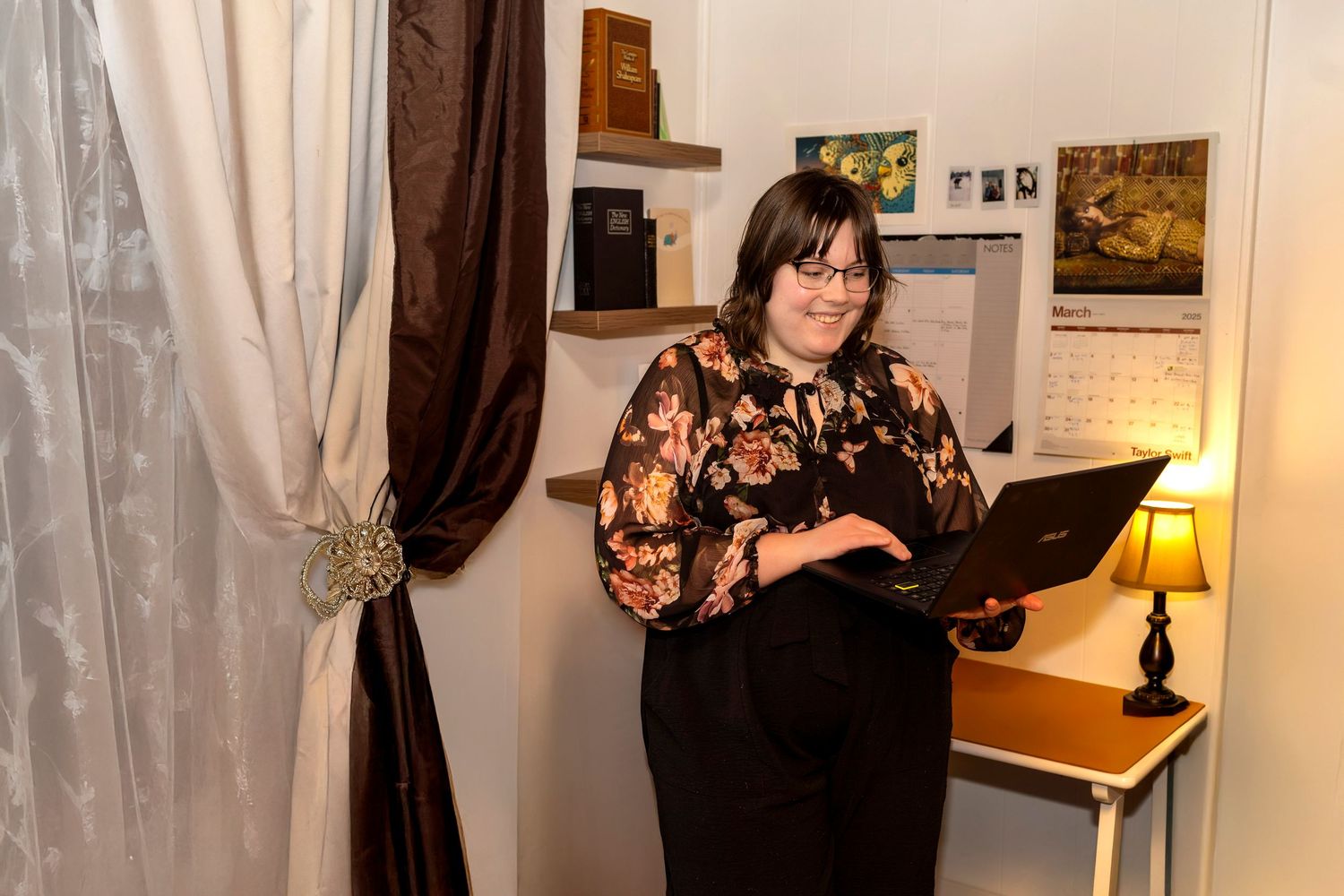 Woman with glasses smiling while holding a laptop in a cozy decorated room with warm lighting and artwork on the walls.