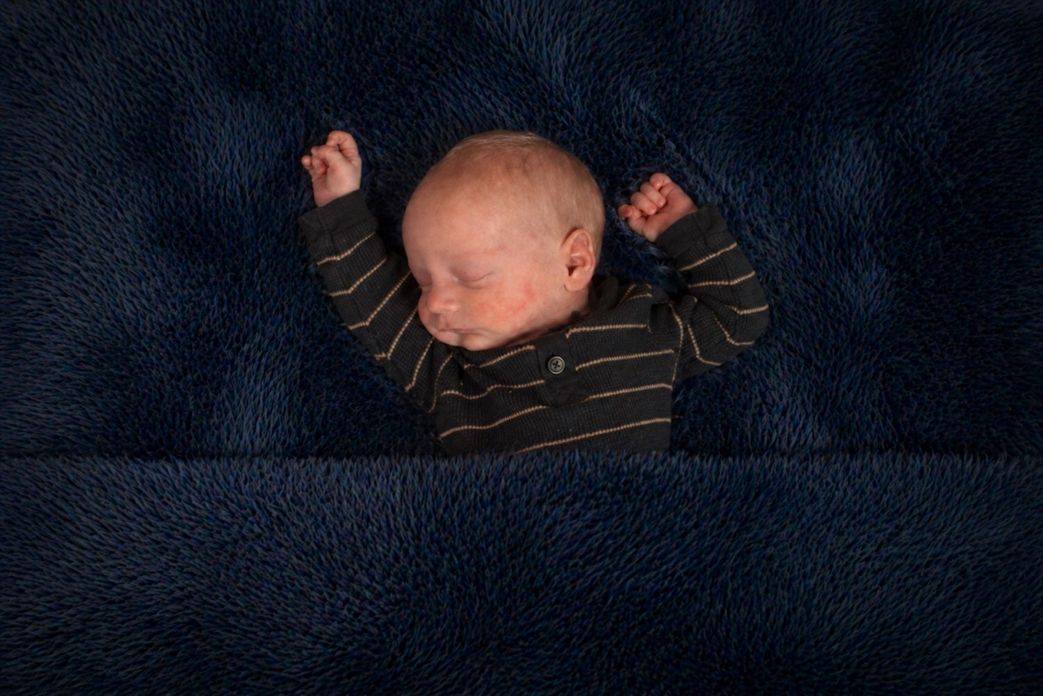 Newborn baby sleeping peacefully on a dark navy blue fluffy blanket wearing a striped onesie with arms raised.