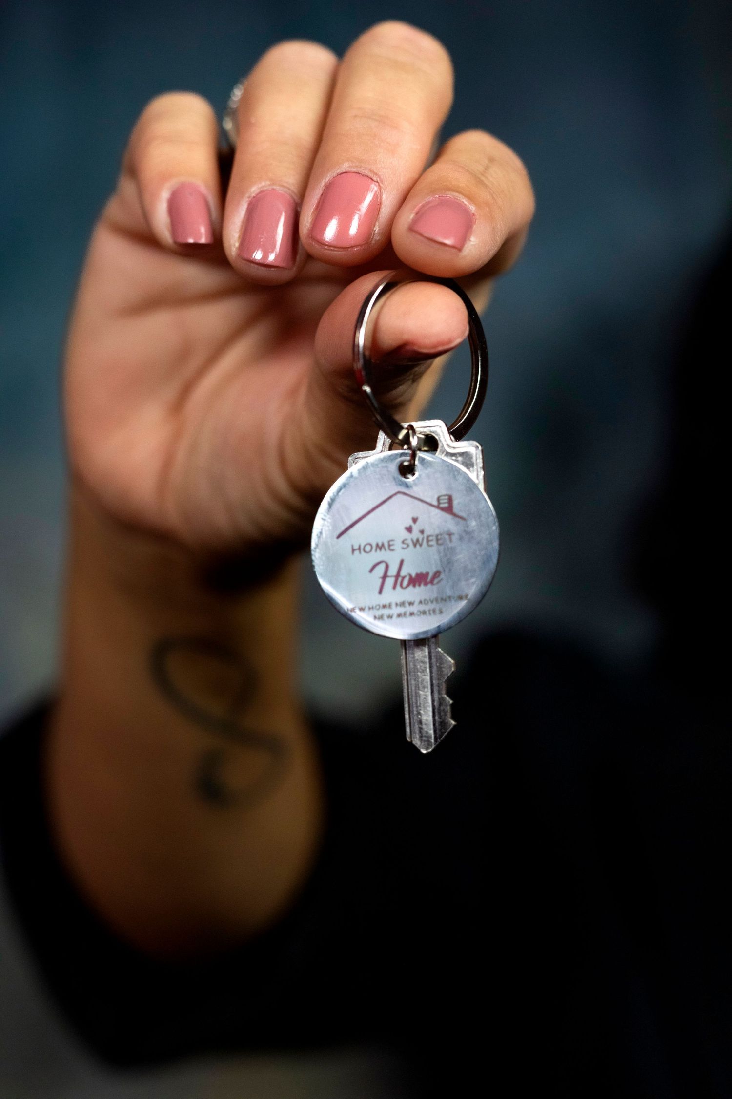 A hand with pink nails and a tattoo holds a house key with a Home Sweet Home keychain.