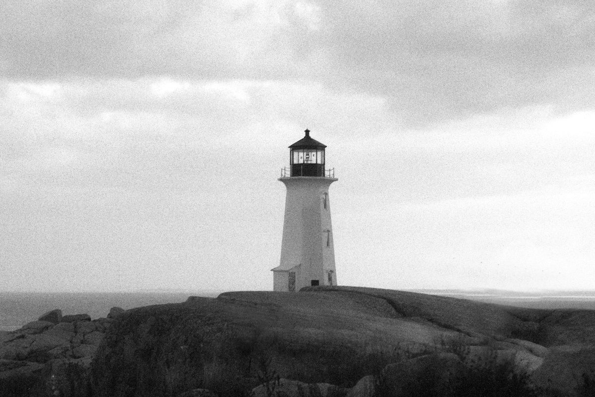 Black and white photo of a classic lighthouse perched on rocky coastal cliffs under a cloudy sky.