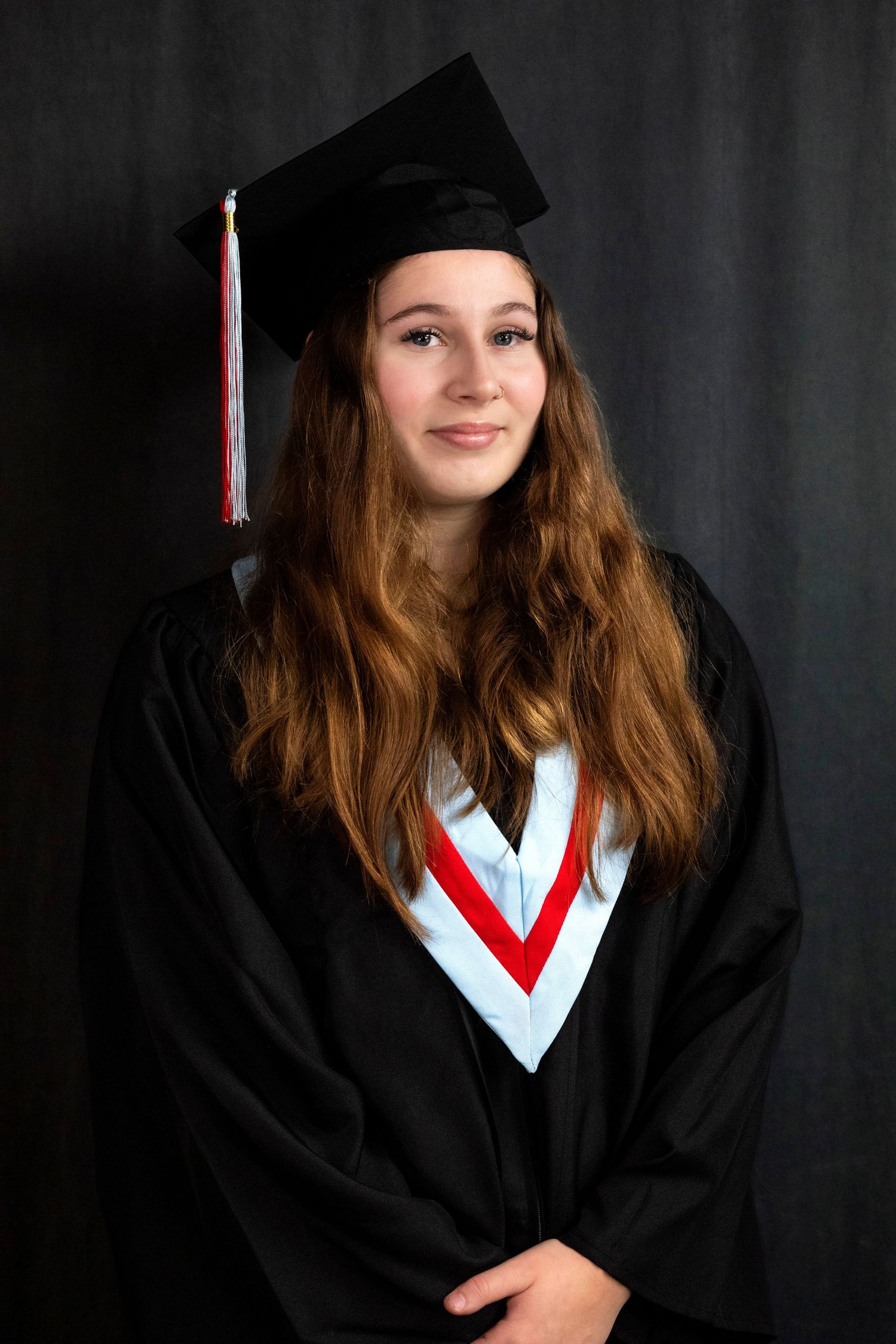 Young woman in black graduation cap and gown with red and white hood, smiling against dark background.