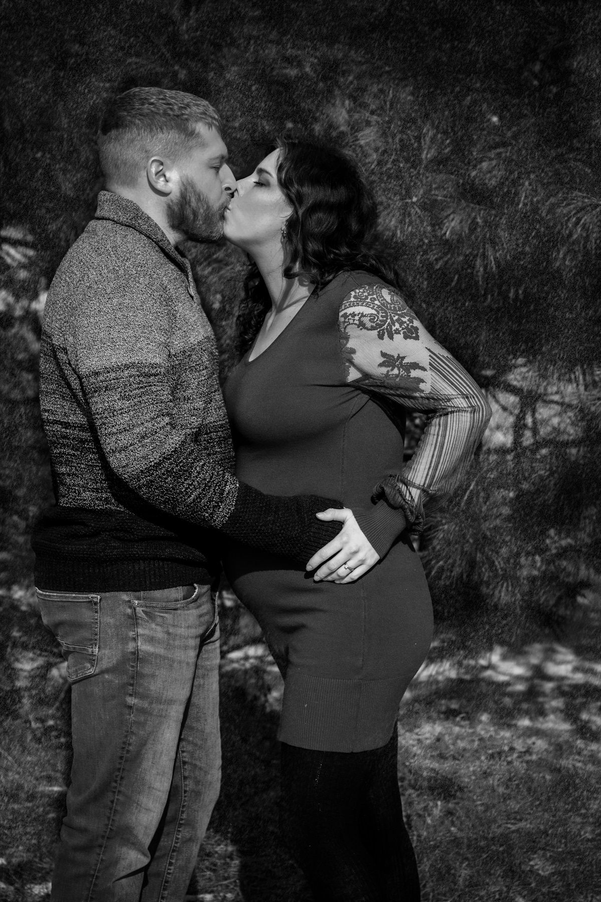 Black and white photo of a pregnant couple kissing outdoors, woman with tattoos in a dress, man in sweater and jeans.