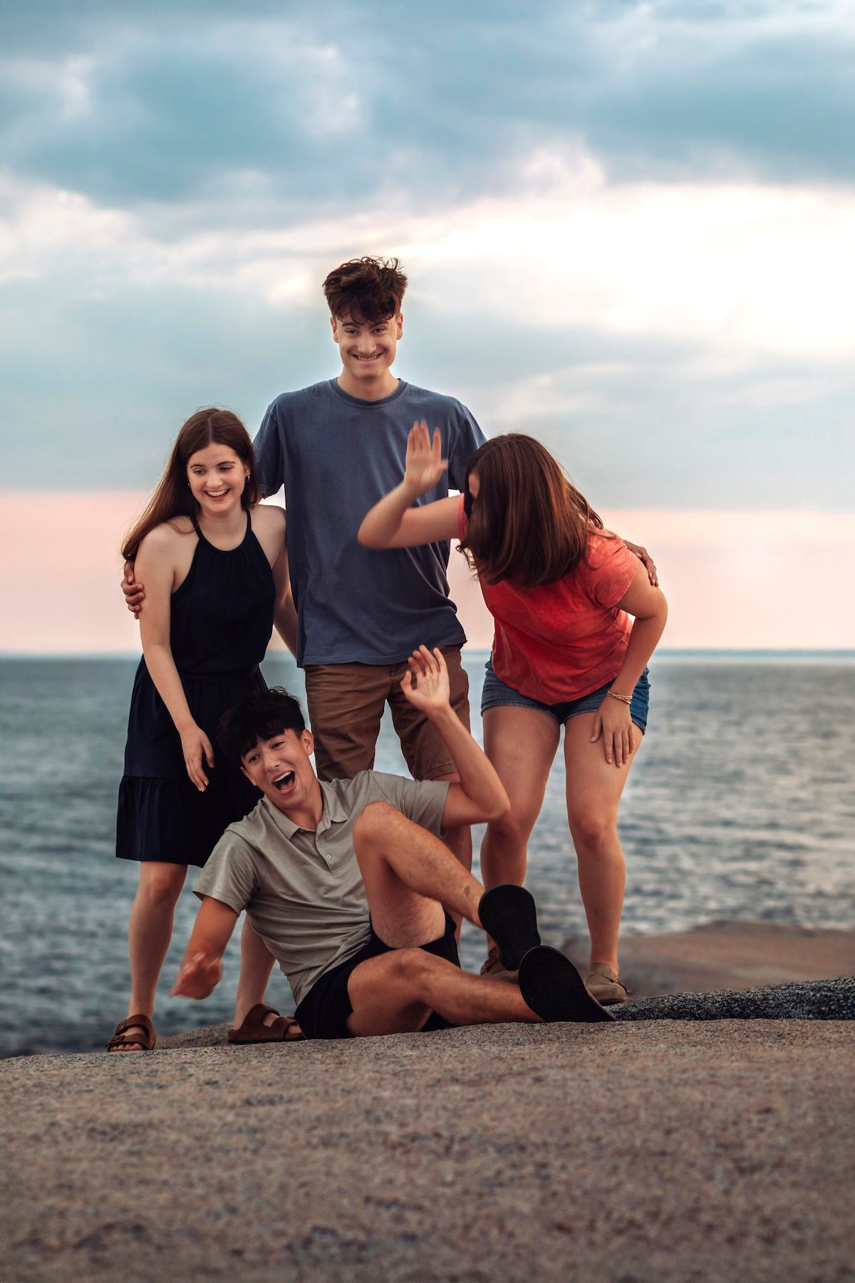 Four young friends laughing and posing together on a seaside pier at sunset, enjoying a fun outdoor gathering.