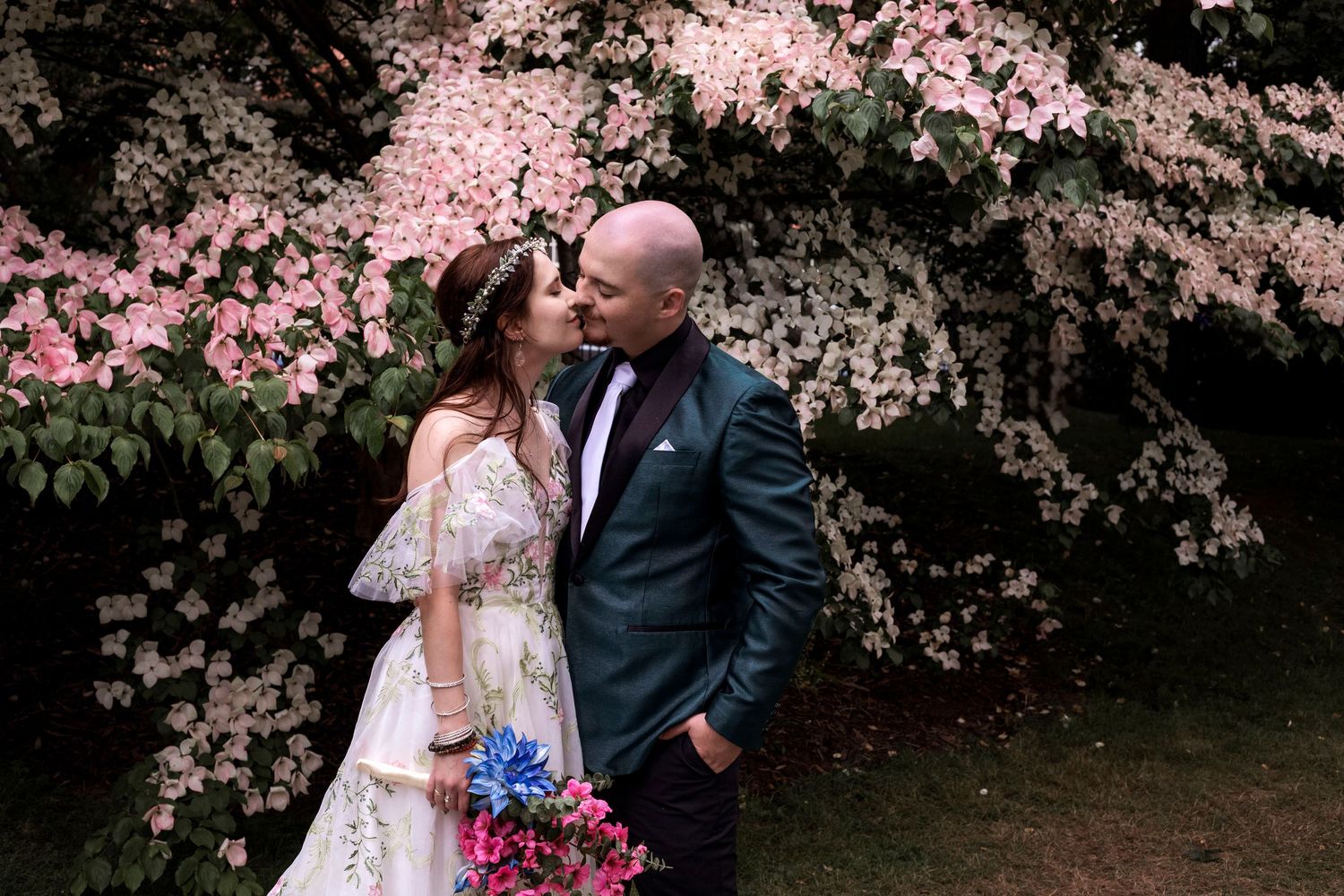 Bride and groom sharing a romantic kiss in front of blooming pink and white dogwood trees, bride holding colorful bouquet.