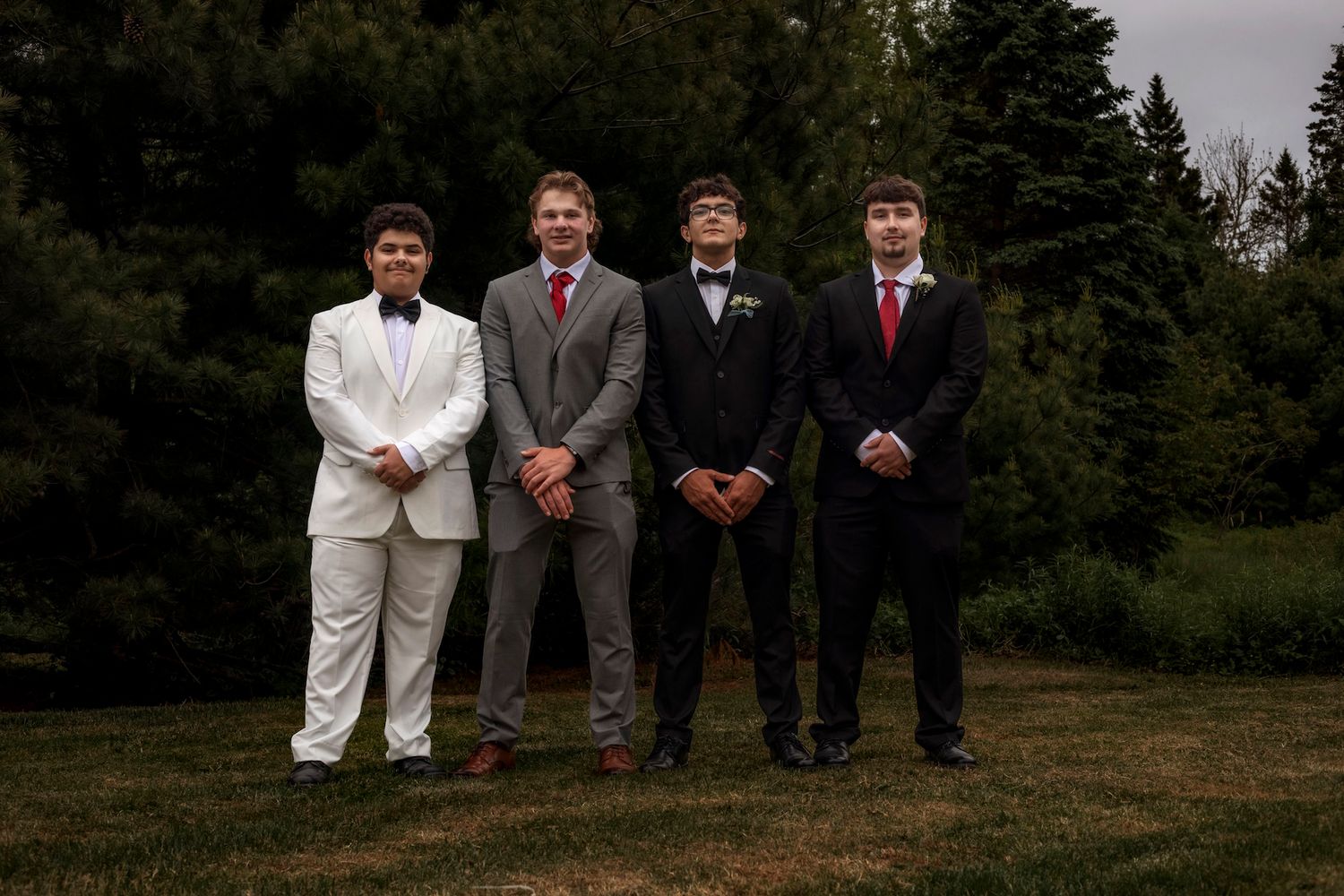 Four young men in formal attire pose outdoors on grass with evergreen trees in the background.