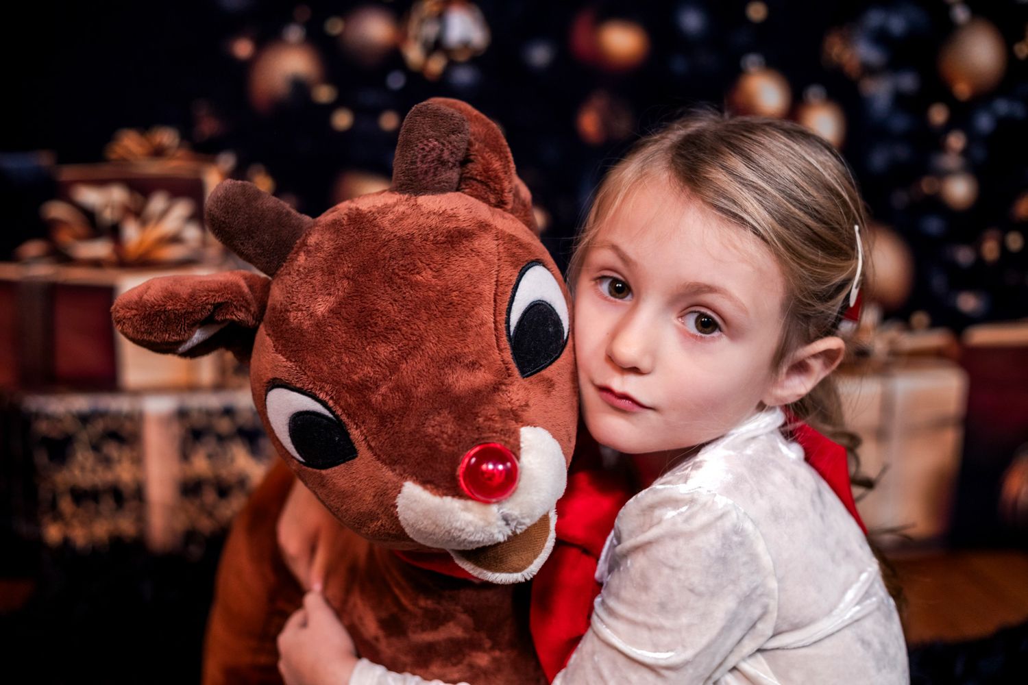Young girl in a white blouse hugs a Rudolph the Red-Nosed Reindeer plush toy in front of a festive Christmas backdrop with gold ornaments.
