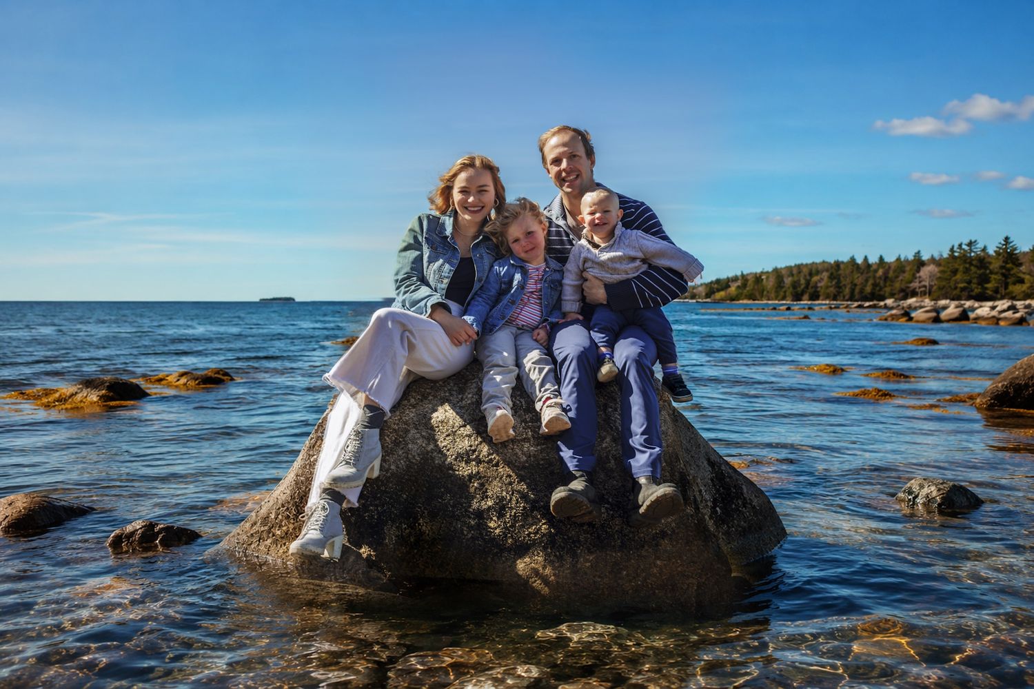 Halls Harbour Family of four smiling on a rocky shoreline with blue water and pine trees in the background on a sunny day.