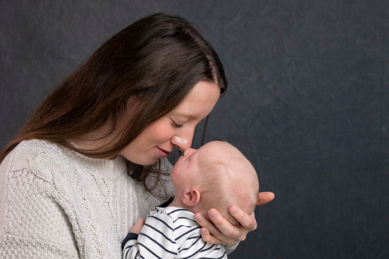 Halifax ns A mother tenderly holds her newborn baby close, touching foreheads in an intimate moment against a dark gray backdrop.