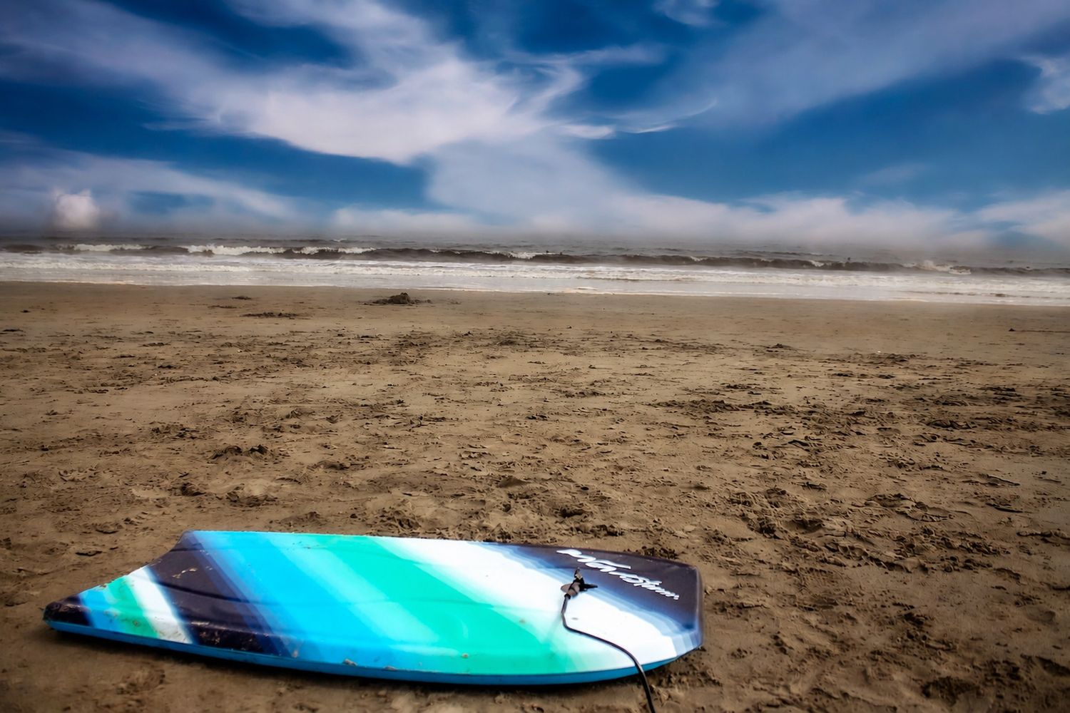 Bridgewater ns beaches, A blue and teal surfboard resting on sandy beach with ocean waves and dramatic cloudy sky in background.