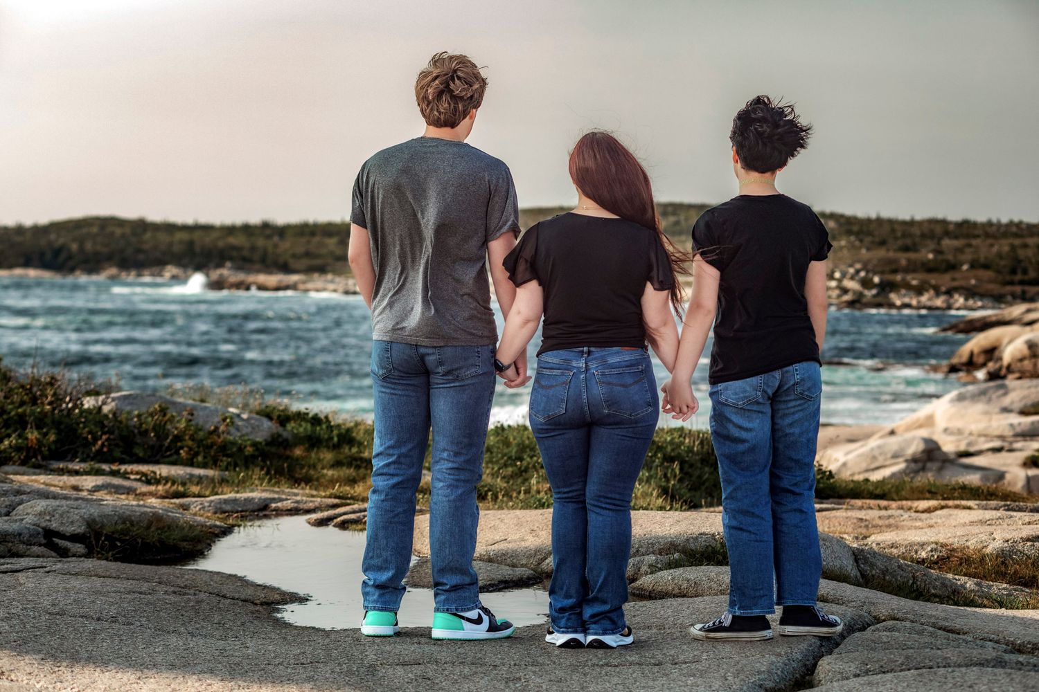 Nova Scotia coast, three people holding hands while standing on rocky coastal shoreline, gazing at the ocean at sunset.