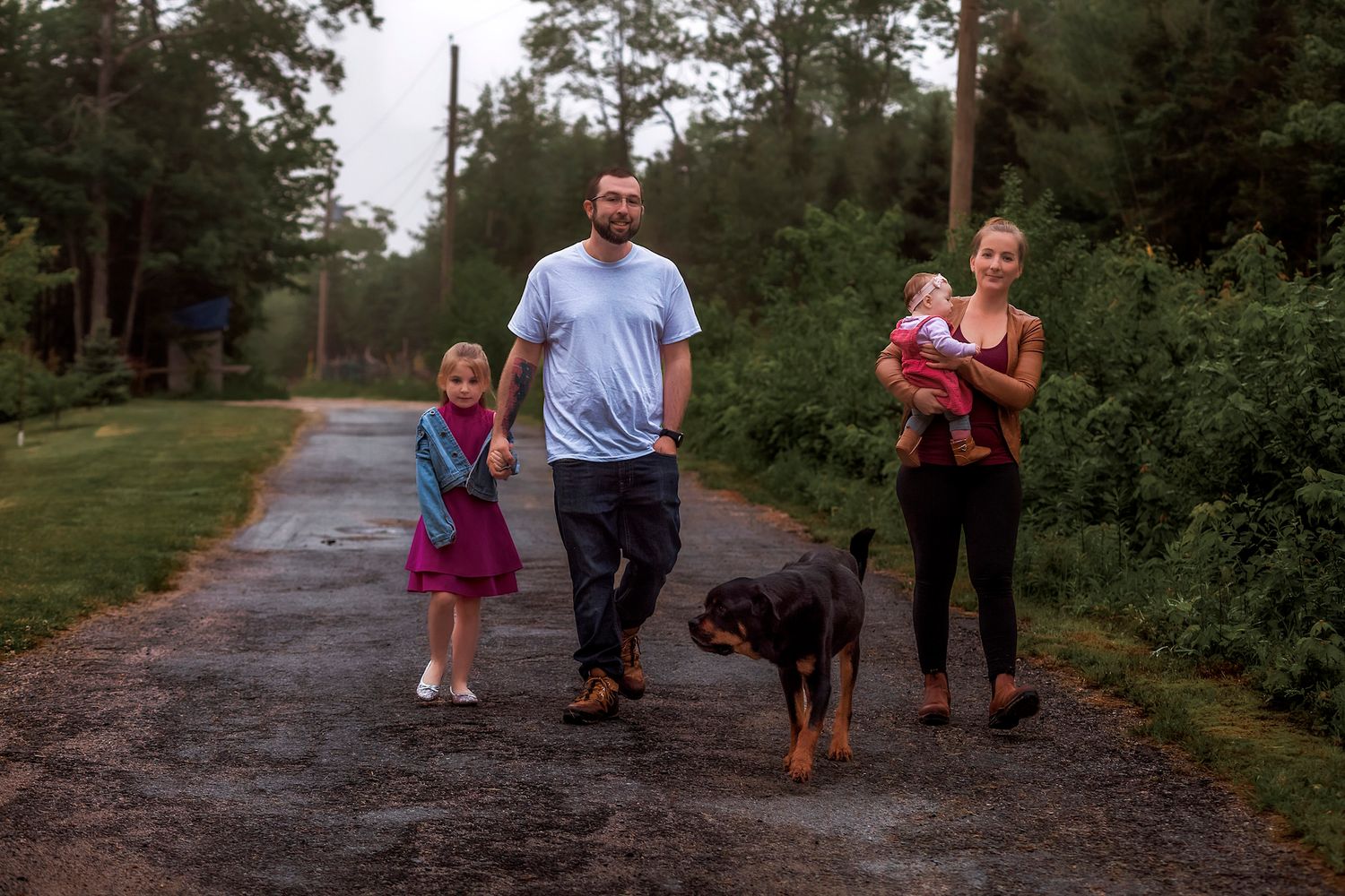 Family with young girl, baby, and Rottweiler walking on a rural gravel path surrounded by lush green trees.