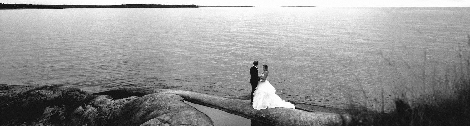 in Halifax a Black and white photo of a bride and groom standing on rocky shoreline by a calm lake on their wedding day.