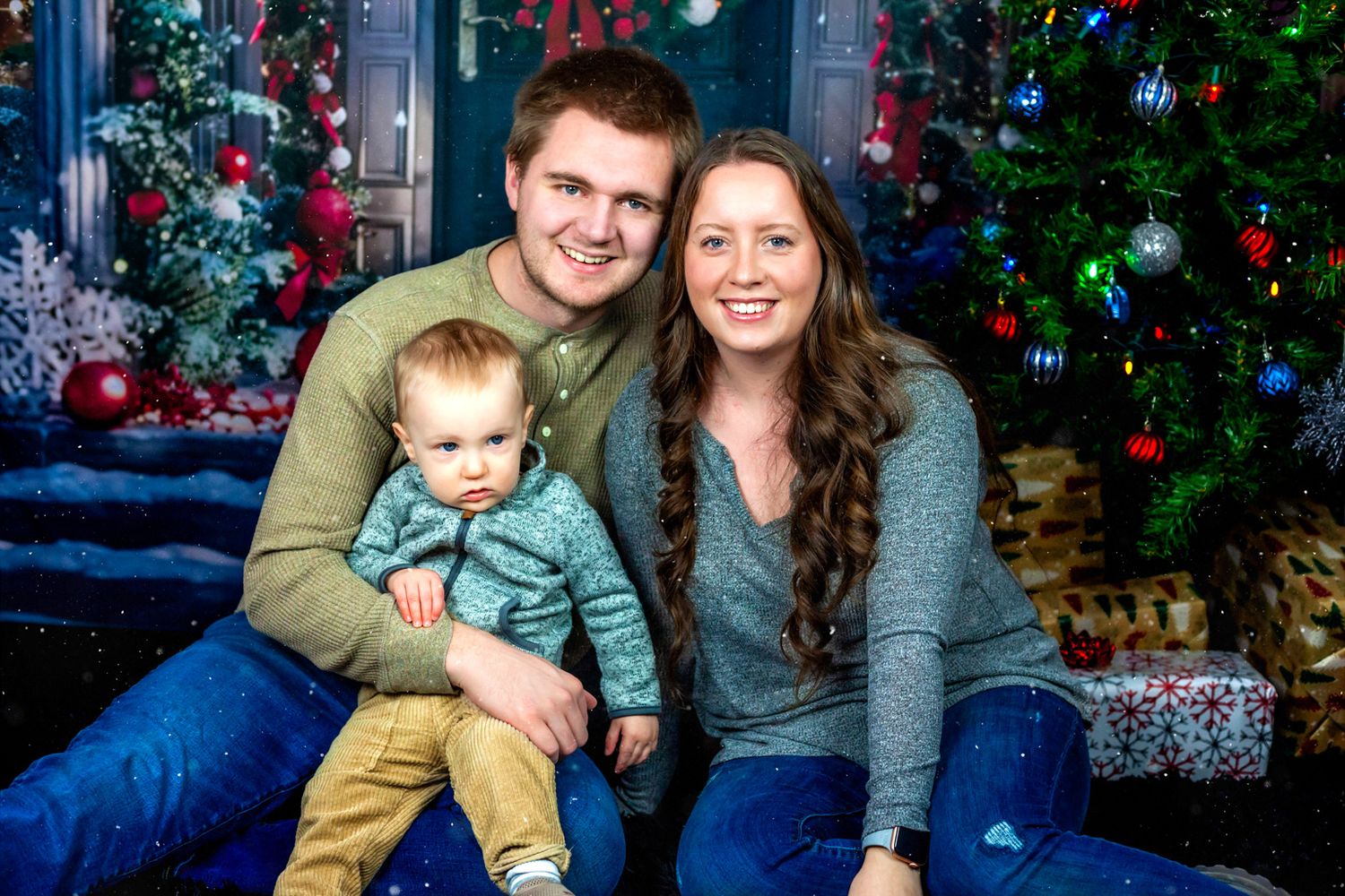 A smiling man and woman sit together holding a toddler in front of decorated Christmas trees and wrapped gifts in a festive family portrait.