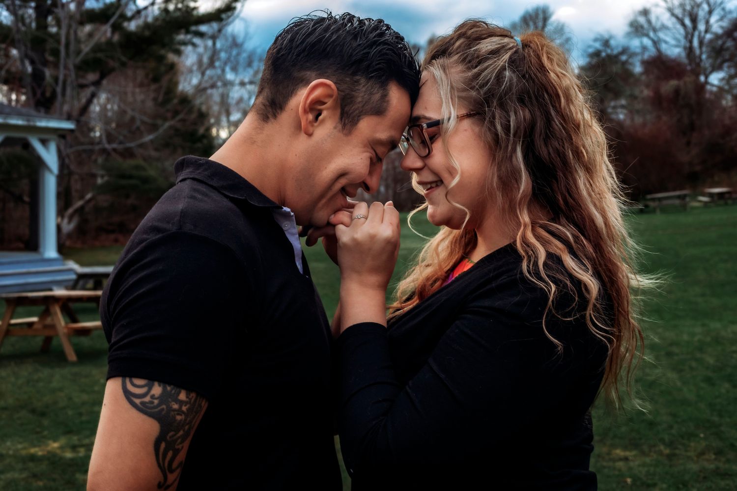 A couple shares a romantic moment outdoors, touching foreheads and smiling, man with tattoos wearing black shirt.