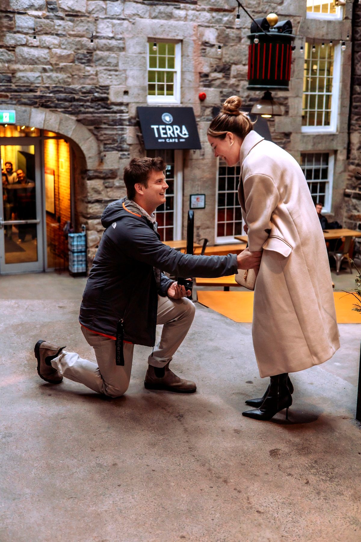 Man proposing on one knee to woman outside Terra Cafe in a stone courtyard, romantic marriage proposal scene.
