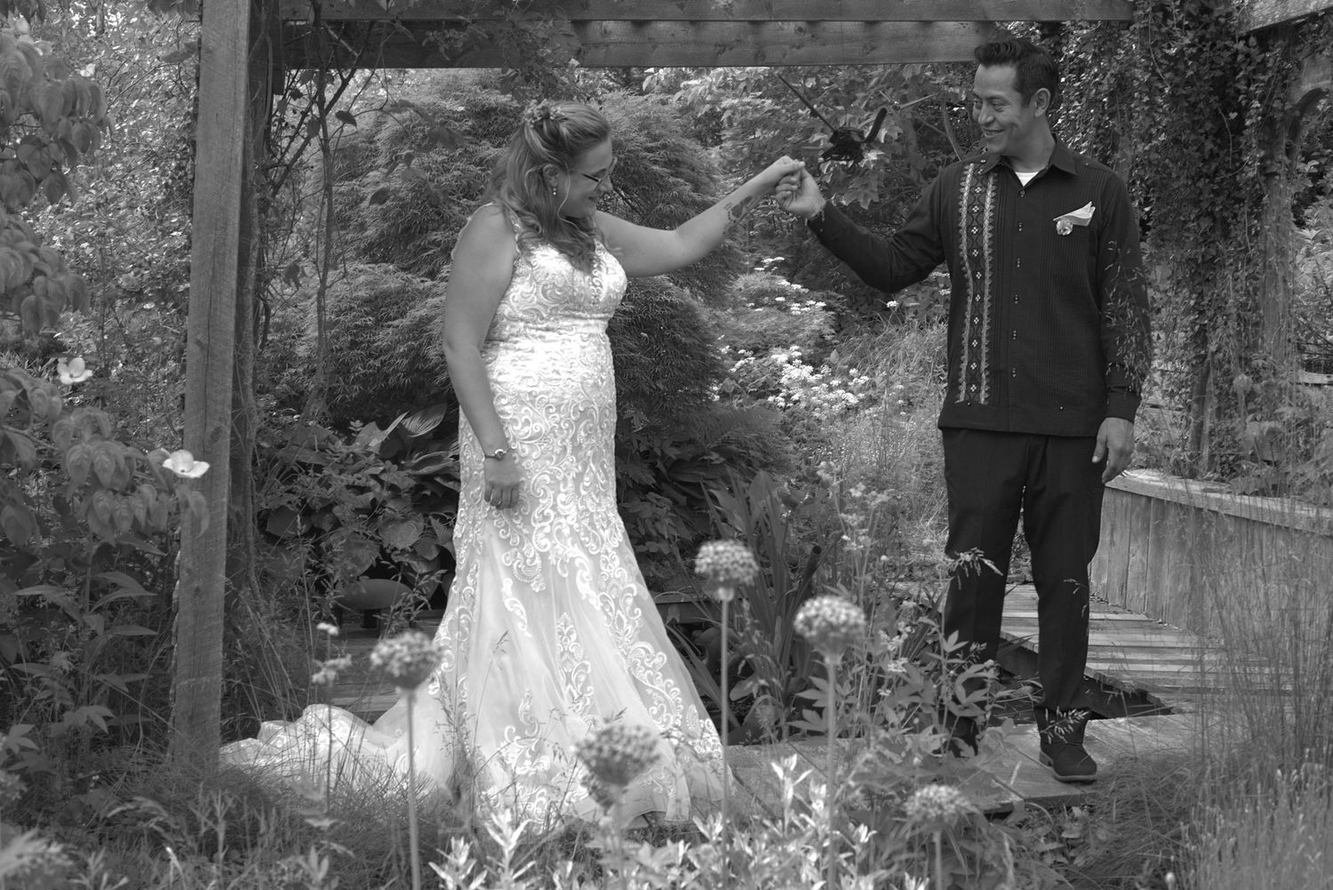 Black and white photo of a bride in lace gown and groom dancing outdoors under a wooden arbor surrounded by lush gardens.