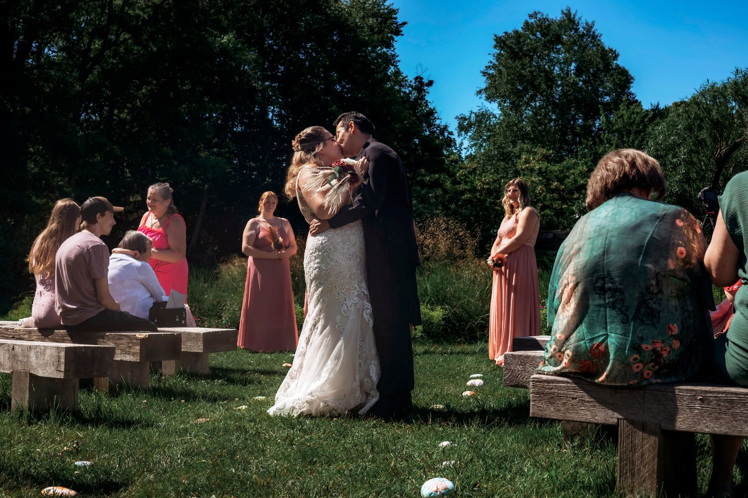 Bride and groom share first kiss at outdoor wedding ceremony with bridesmaids in pink dresses and guests seated on wooden benches.