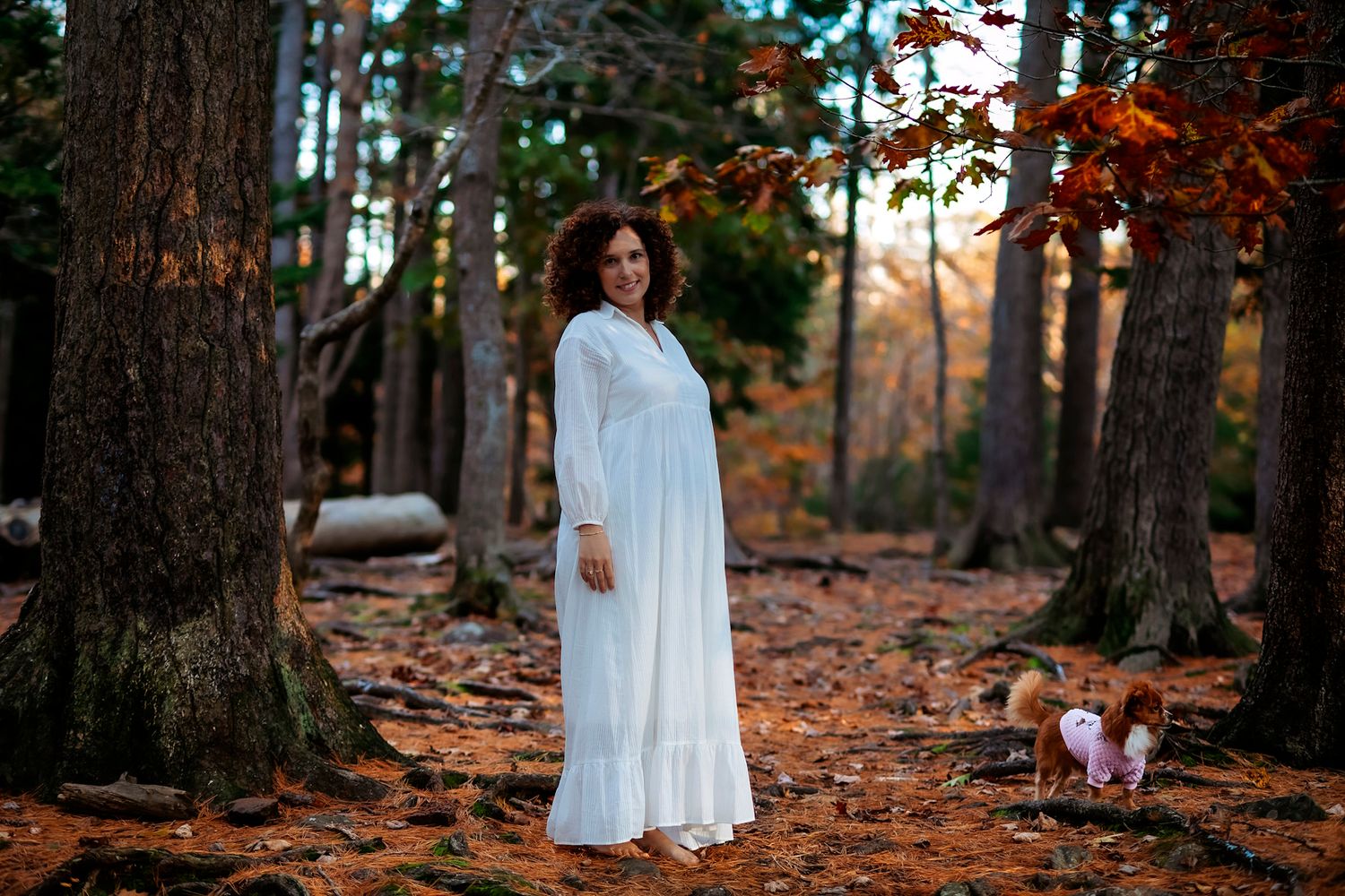 Woman in white dress standing among autumn trees with small dog in colorful fall forest setting.