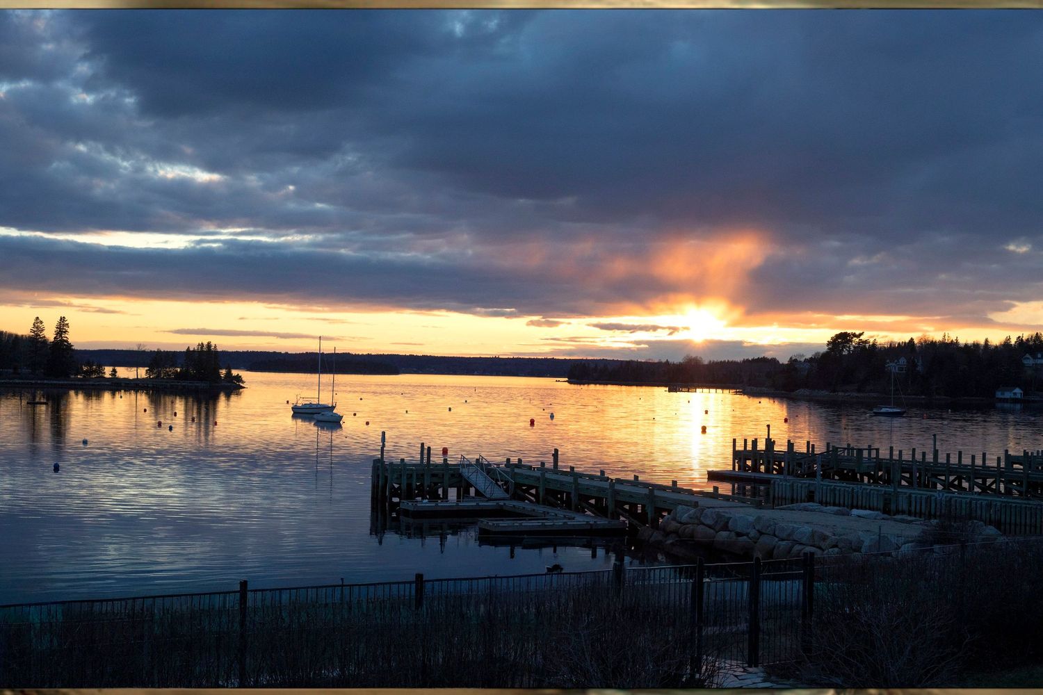 Stunning sunset over a calm harbor with docks and boats reflecting golden light on the water under dramatic cloudy skies.