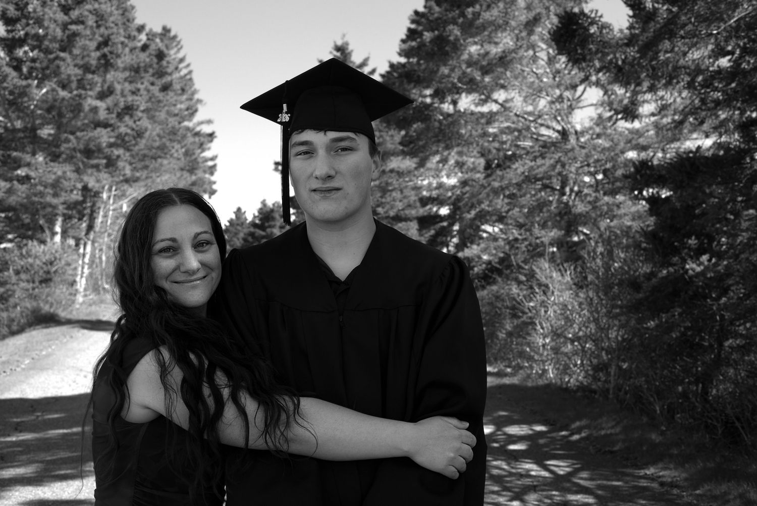 Black and white photo of a couple, man in graduation cap and gown, posing at Graves Island Provincial park near trees.