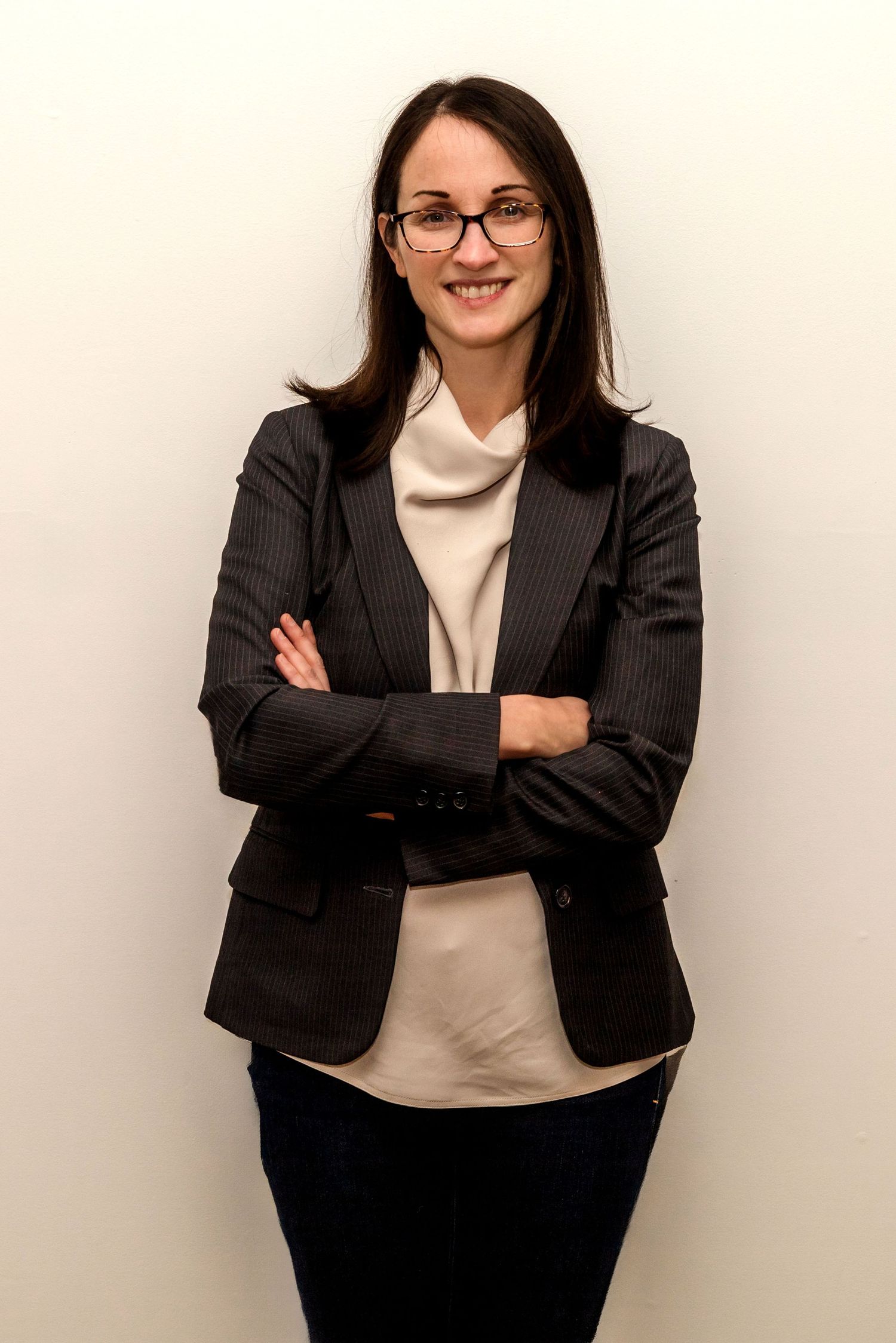 Professional woman in dark blazer and glasses smiling with arms crossed against white background.