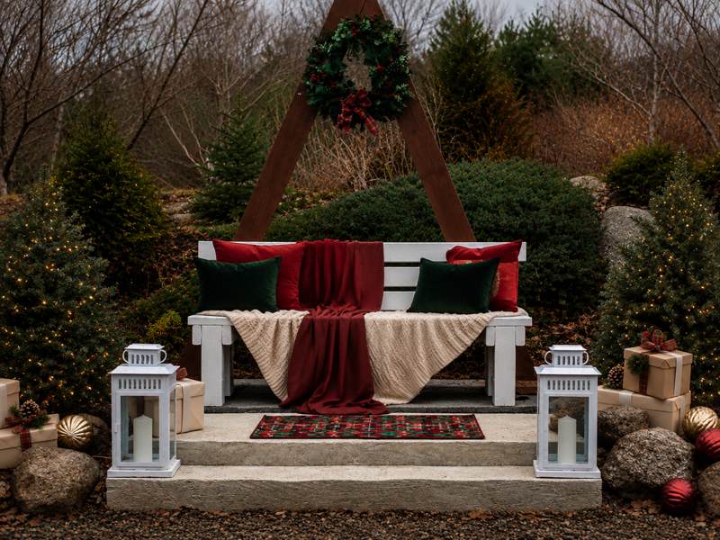Outdoor Christmas photo setup with a white bench, red and green pillows, plaid blankets, wreath on a wooden frame, lanterns, and gifts.