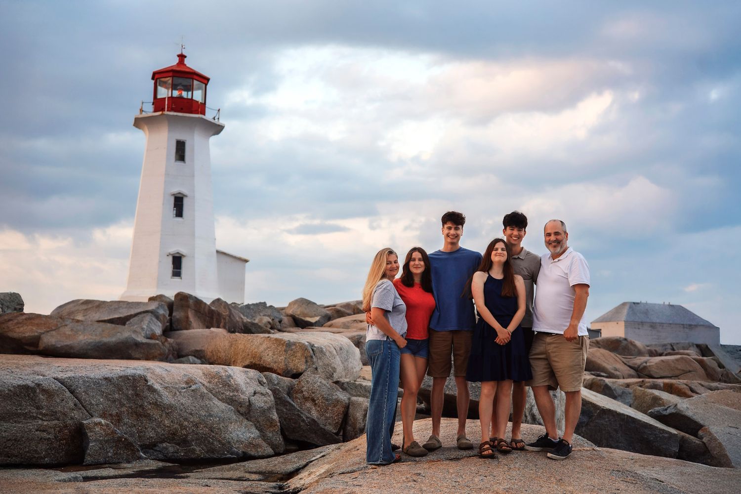 Family of five posing on rocky shore near Peggy's Cove lighthouse under dramatic cloudy sky.