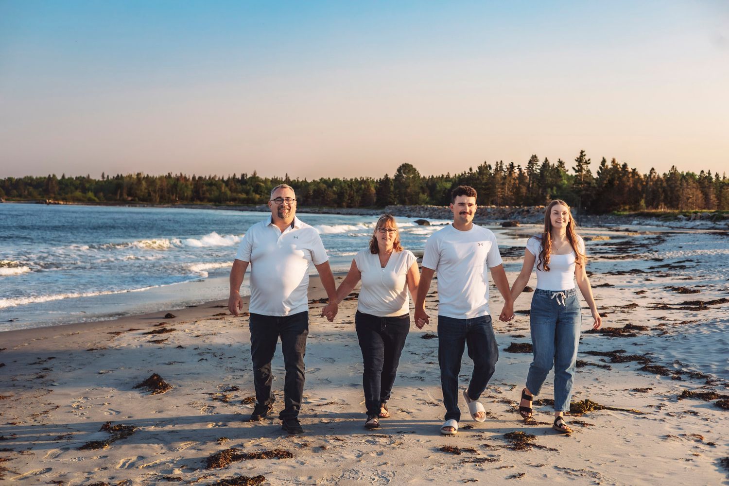 Family of four walking hand-in-hand on a sandy beach at sunset with trees and ocean waves in the background.