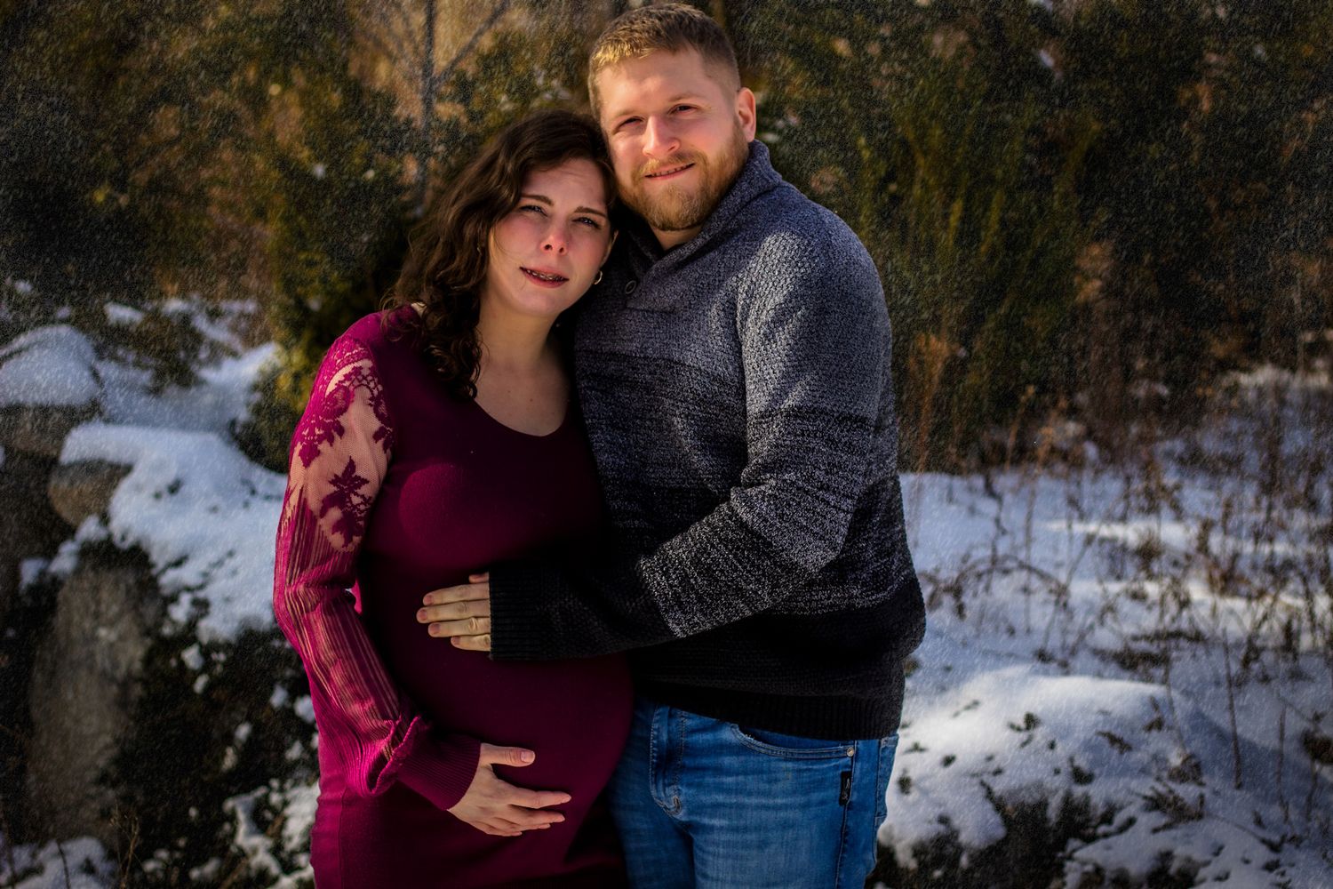 Pregnant couple embracing outdoors in winter snow, woman in burgundy maternity dress, man in gray sweater.