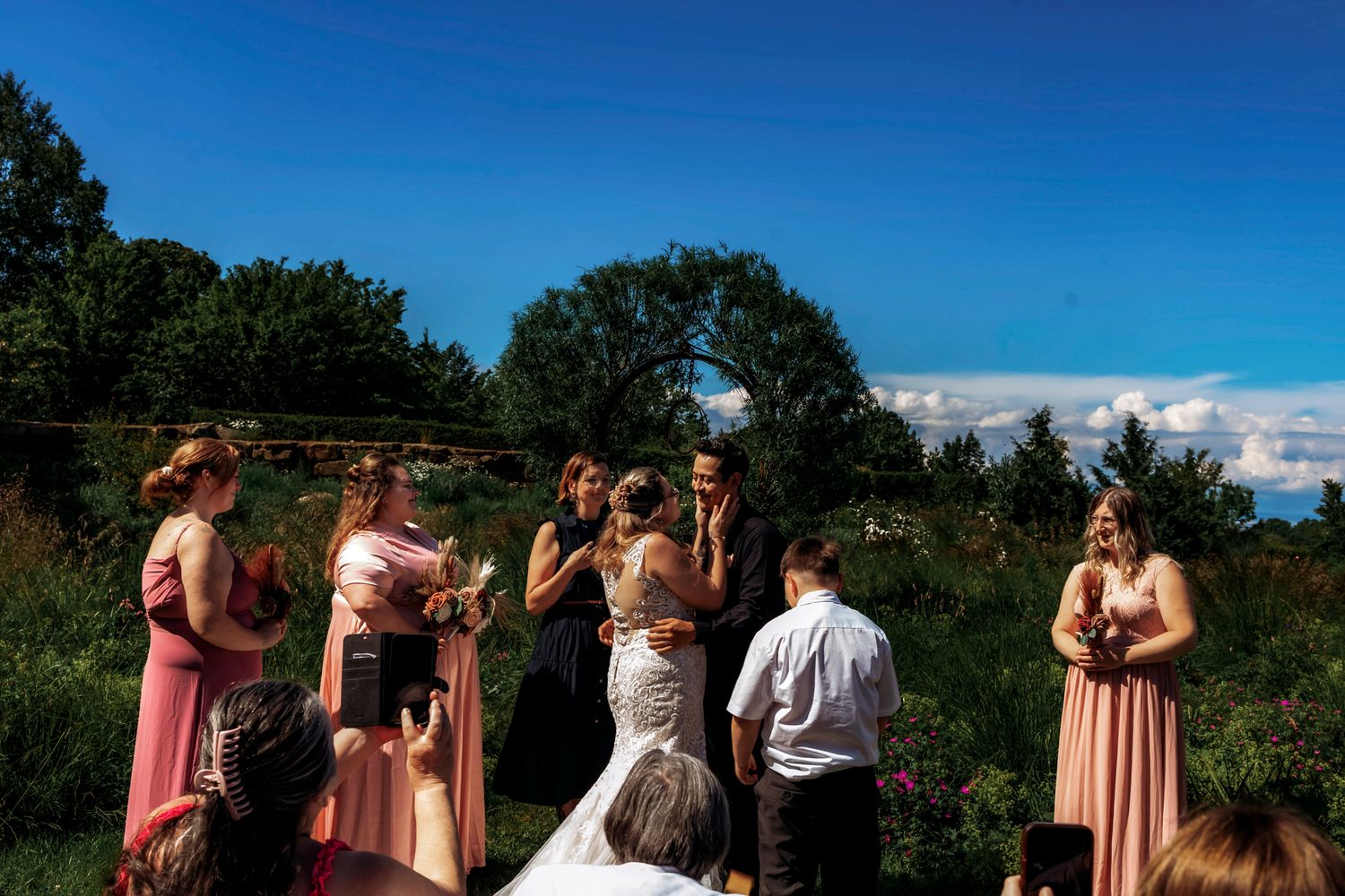 Outdoor wedding ceremony with bridesmaids in pink dresses under blue sky surrounded by lush greenery.