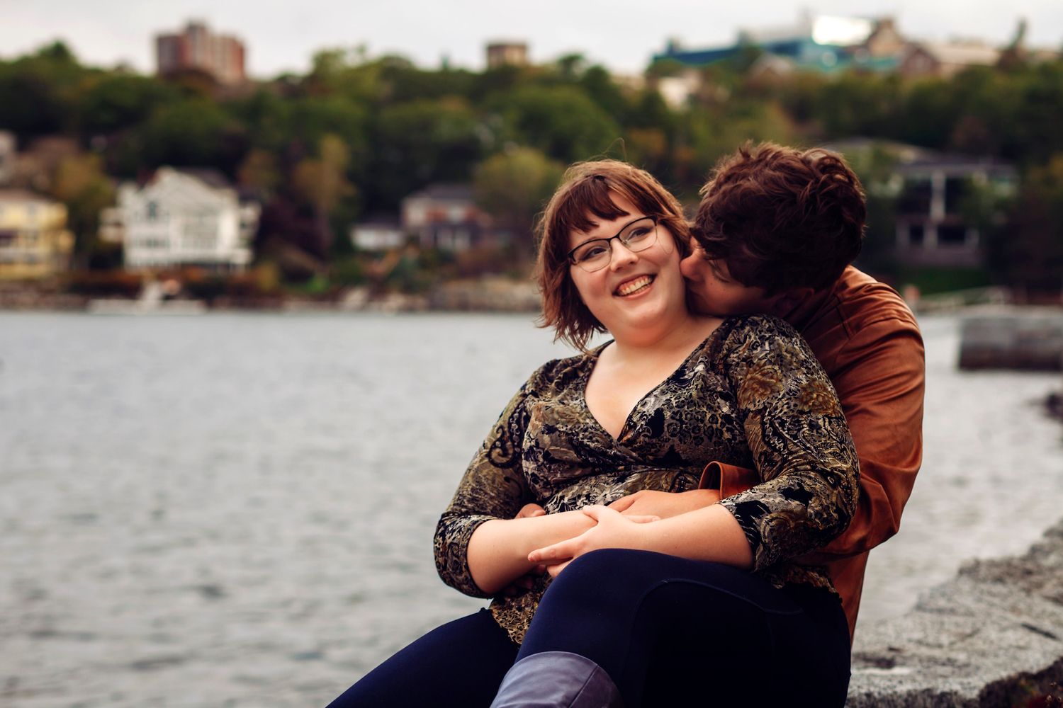 Couple sharing a romantic moment by the waterfront, woman smiling as man kisses her cheek outdoors.