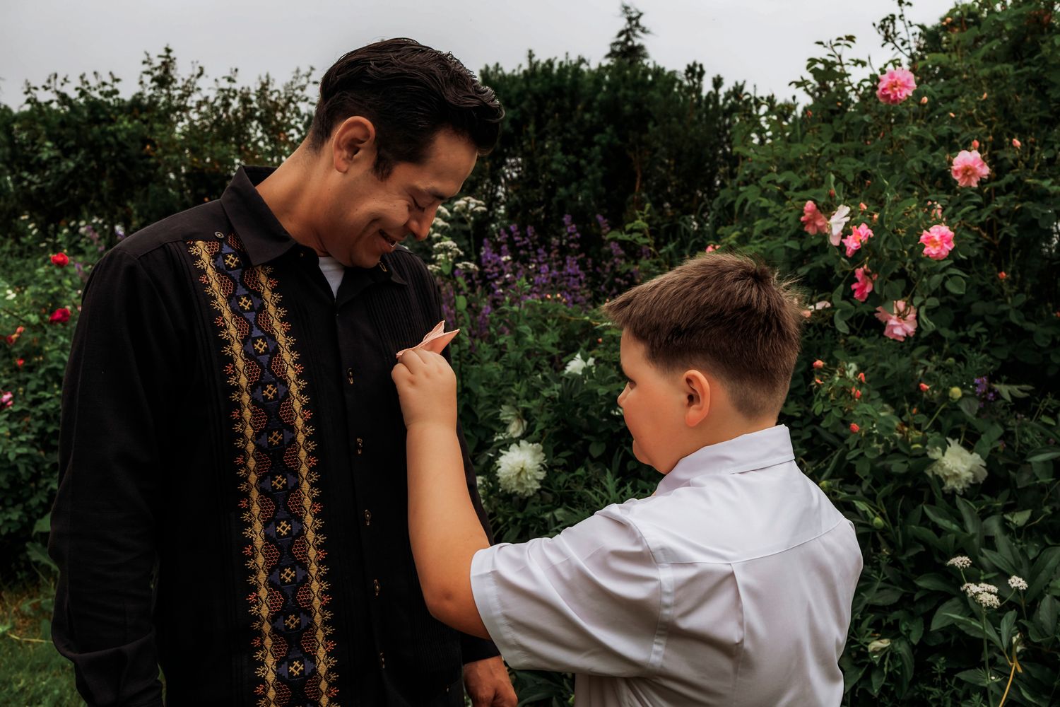 A boy pins a flower to a man's black embroidered shirt in a lush garden with pink roses blooming in the background.